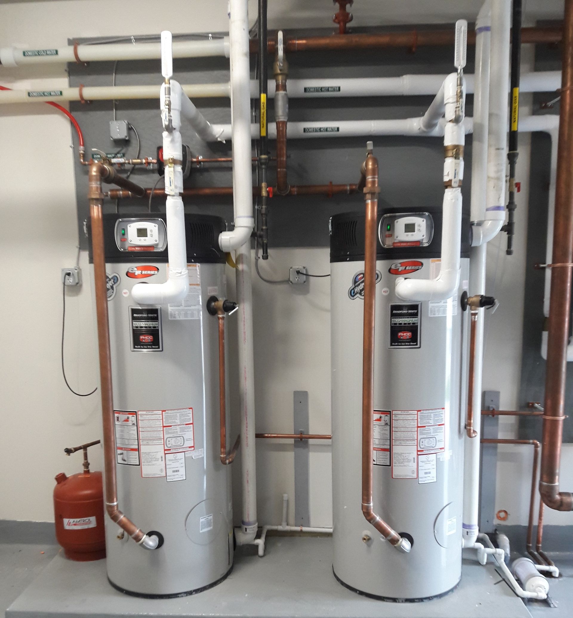 Two large silver water heaters with piping and valves in a utility room