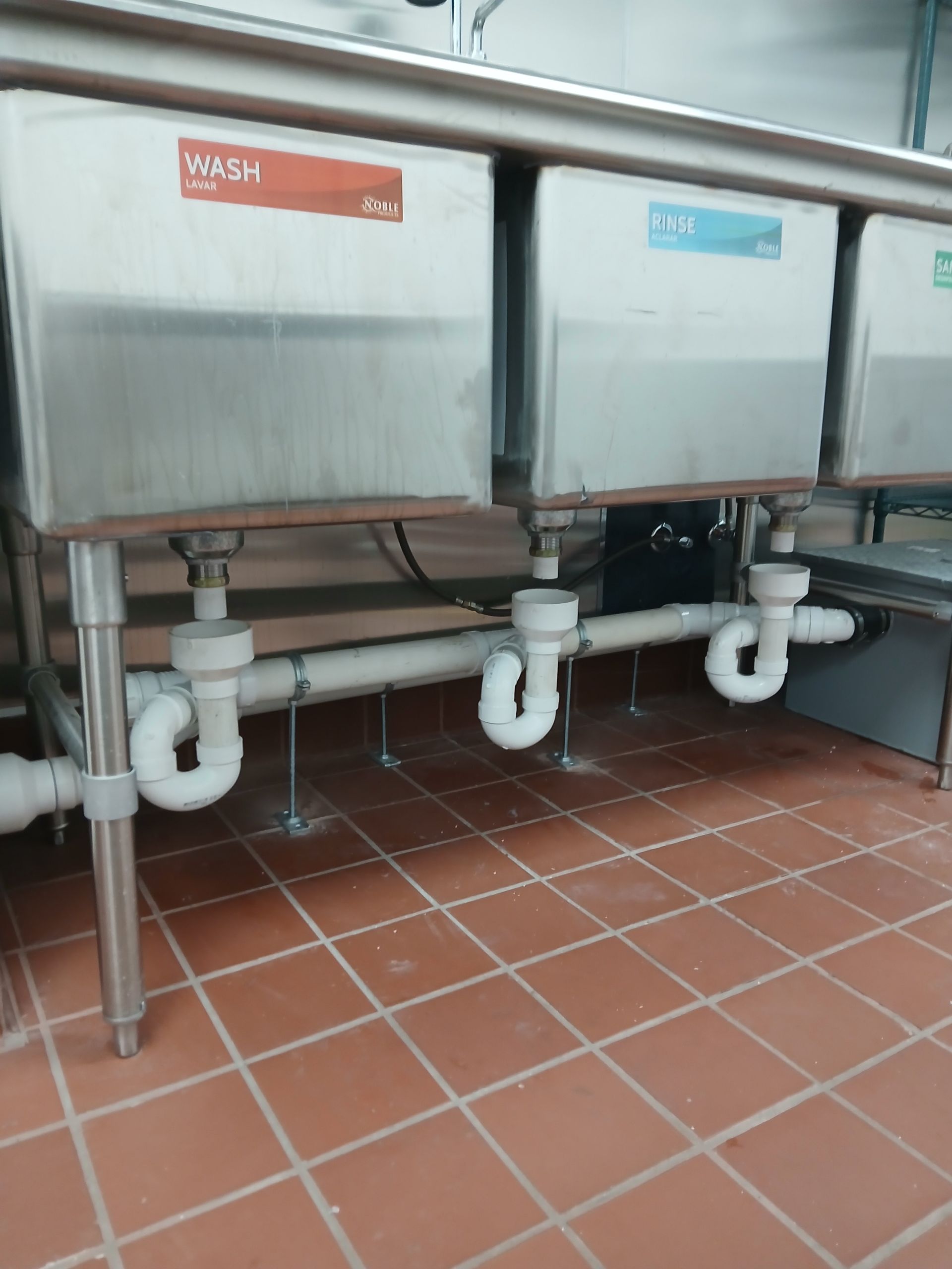 Stainless steel sinks with white plumbing pipes under a tiled counter in a commercial kitchen