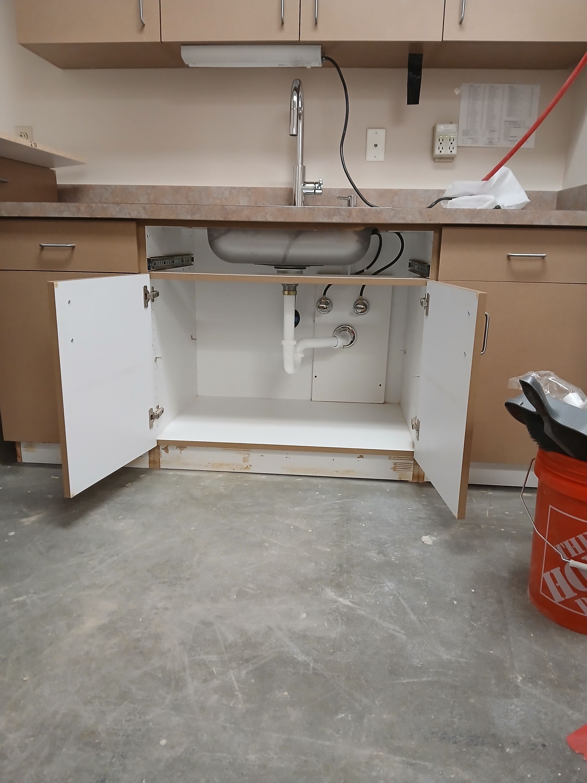 Open under-sink cabinet in a workshop, with plumbing exposed and cabinet doors open.