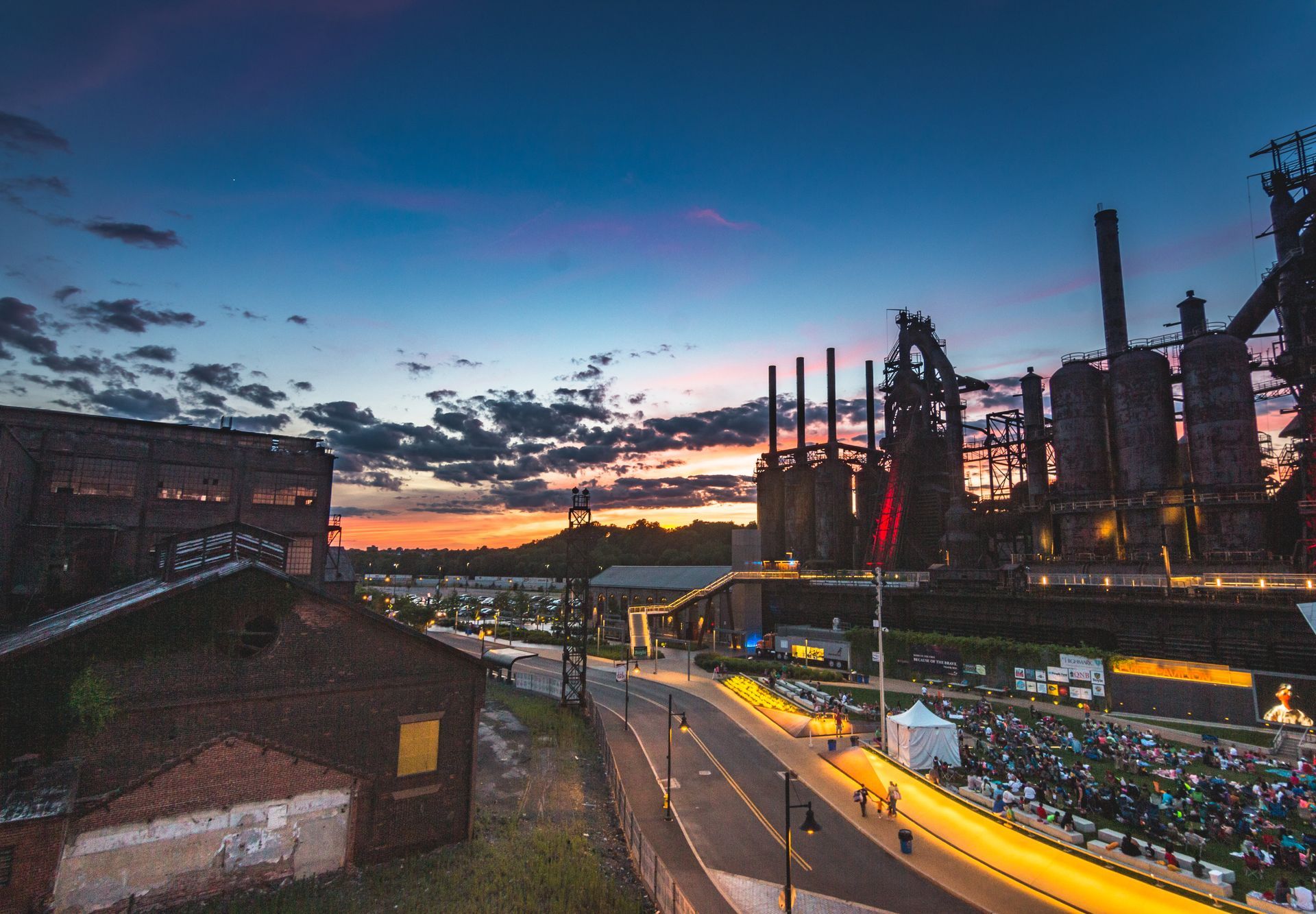 Twilight view of an industrial steel plant with smokestacks, glowing furnace lights, and a rail line in front