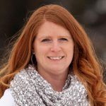 Smiling woman with long red hair wearing a white and grey patterned scarf against a blurred outdoor background.