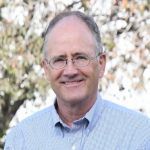 A middle-aged person wearing glasses and a light blue button-down shirt, smiling in front of a tree with blurred leaves.