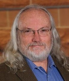 A person with long gray hair and a beard, wearing glasses, a blue collared shirt, and a brown blazer against a brick wall.