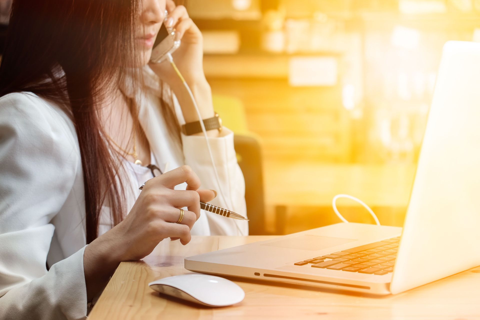 A person in a white blazer talking on a phone while holding a pen near an open laptop on a wooden desk.