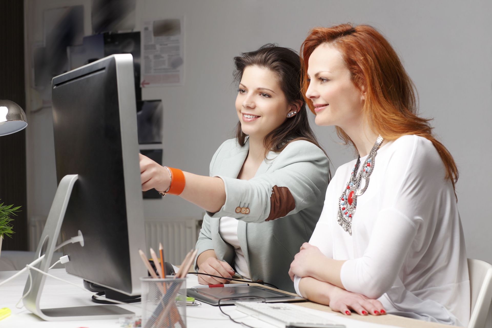 Two colleagues sit at a desk, looking at a computer screen as one points to something on the display.