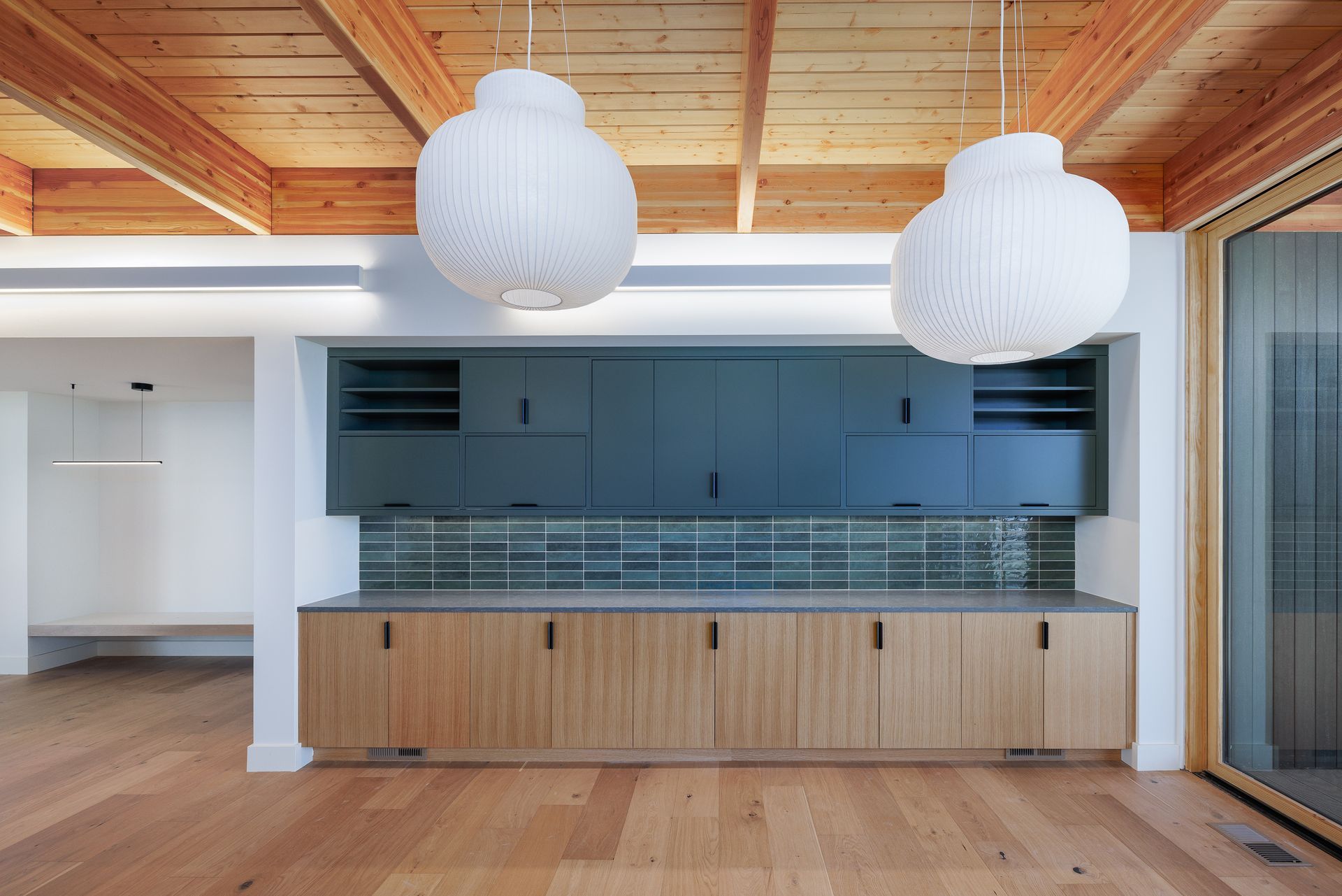 Modern kitchen with wood cabinetry, blue backsplash, white pendant lights, and exposed wood ceiling