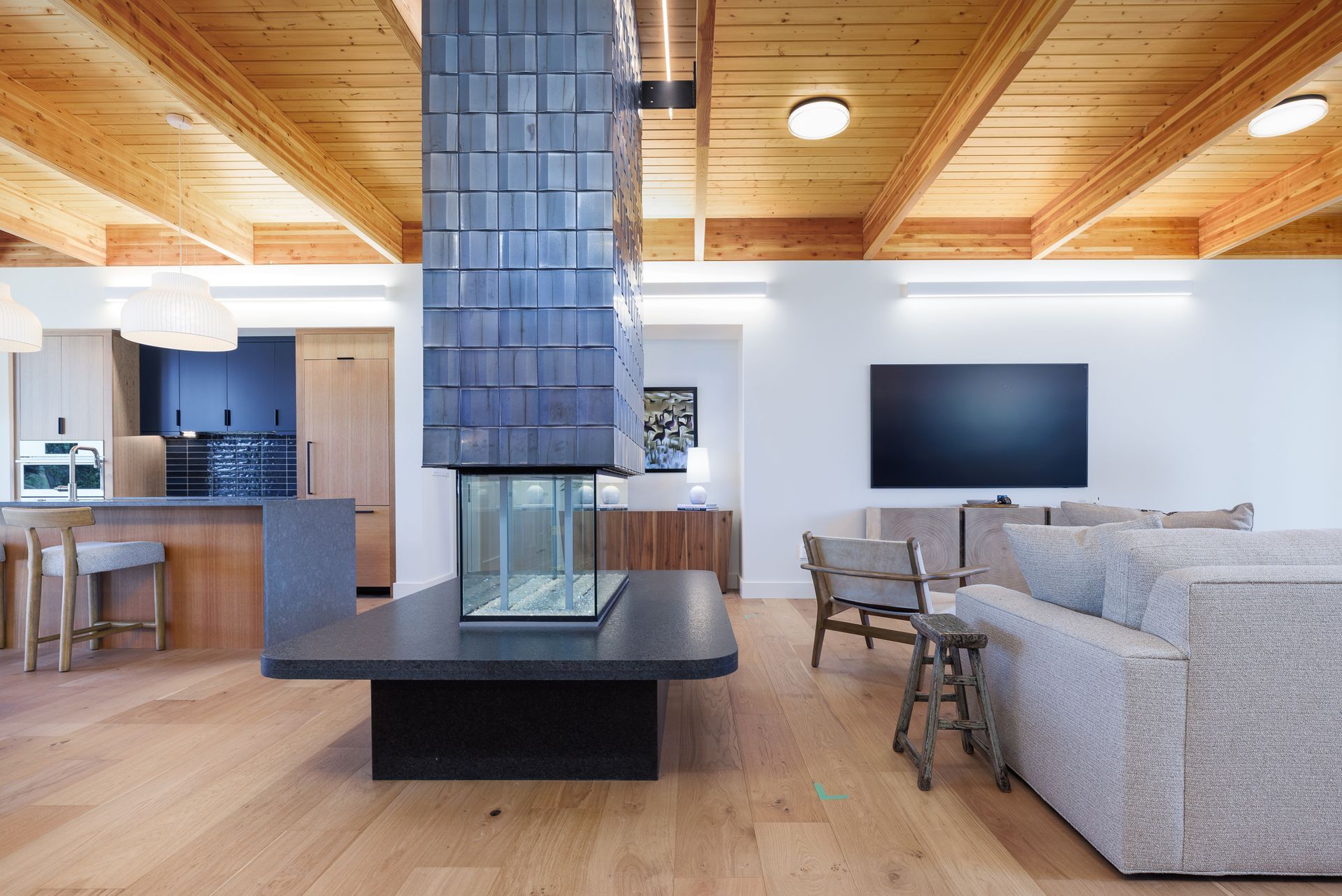 Modern open-plan living room with central fireplace, wood ceiling, sectional sofa, and TV.