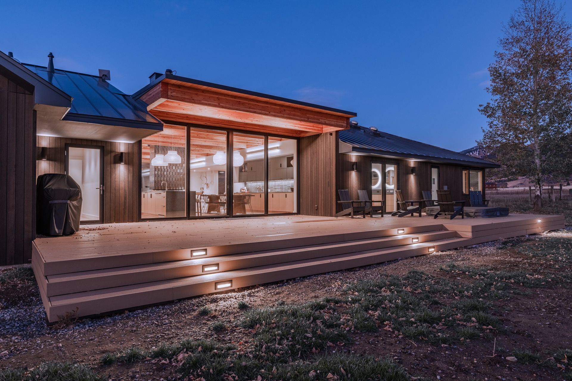 Modern house exterior at dusk with warm lit windows, wooden deck steps, and a blue evening sky