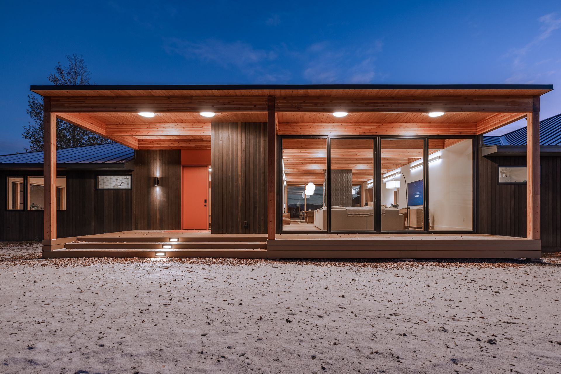 Modern single-story house with warm wood siding and large glass windows at dusk on a snowy lot