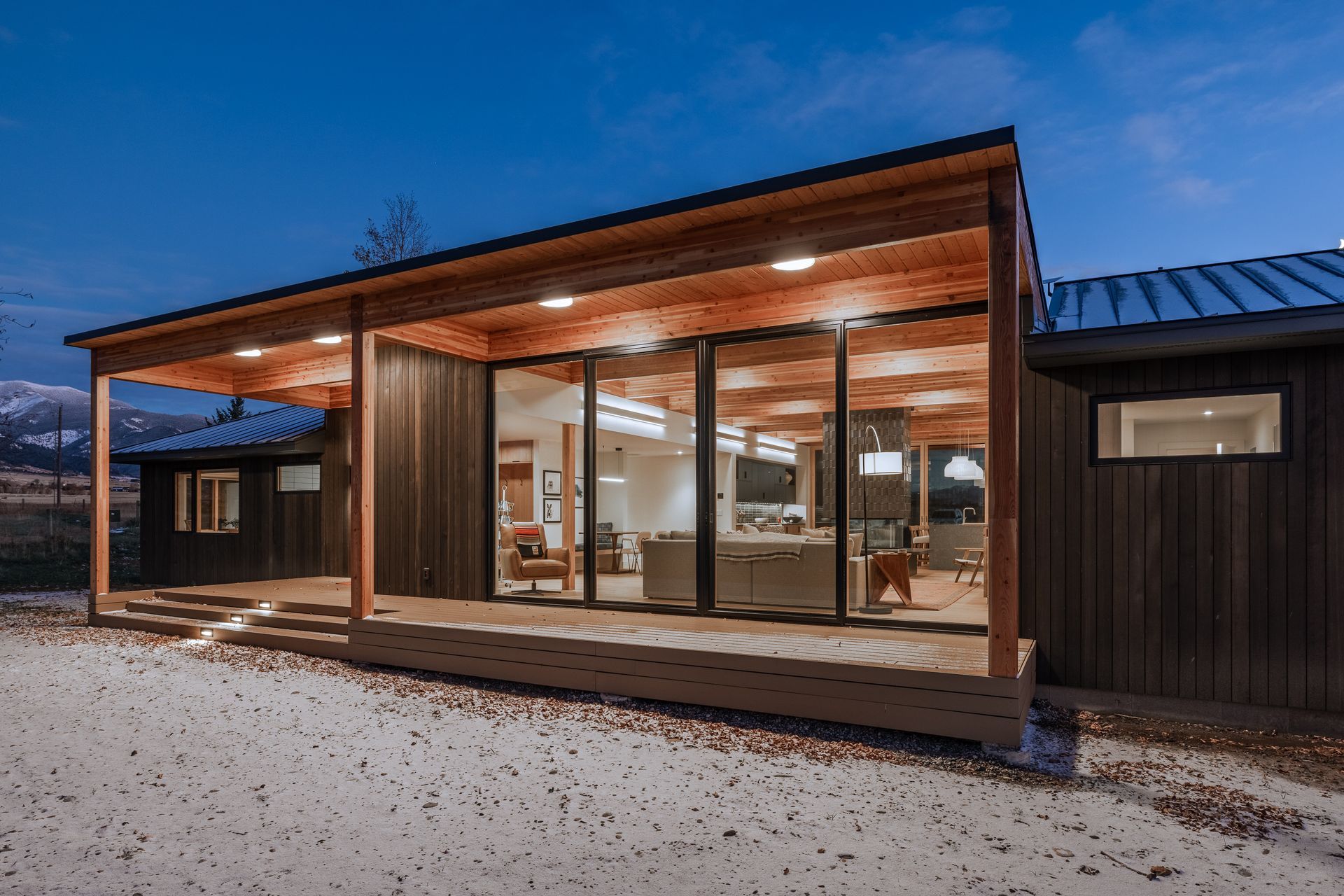 Modern cabin exterior with large glass windows, warm wood deck, and snowy ground at dusk