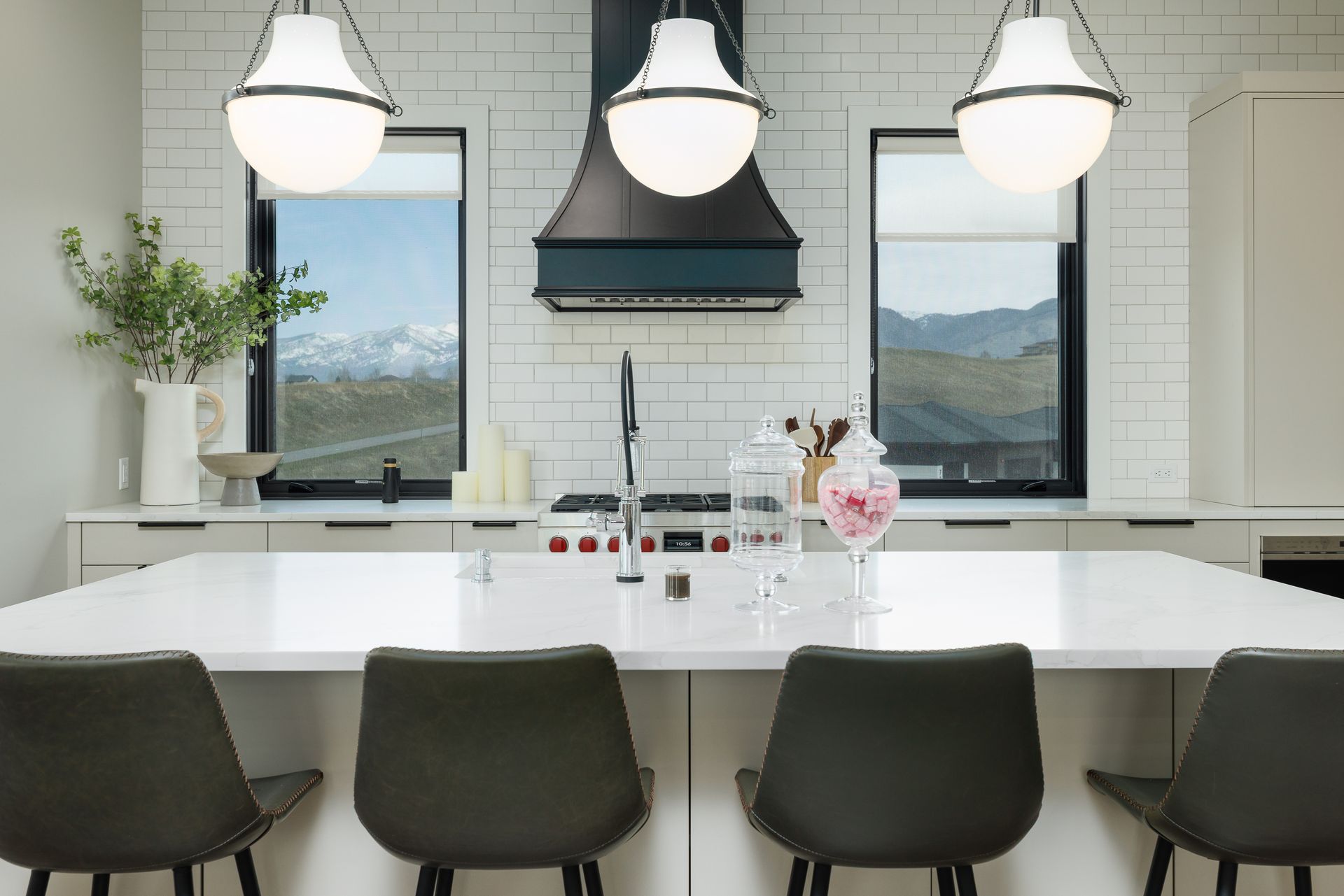 Modern white kitchen with island, three pendant lights, dark bar stools, and large windows overlooking mountains