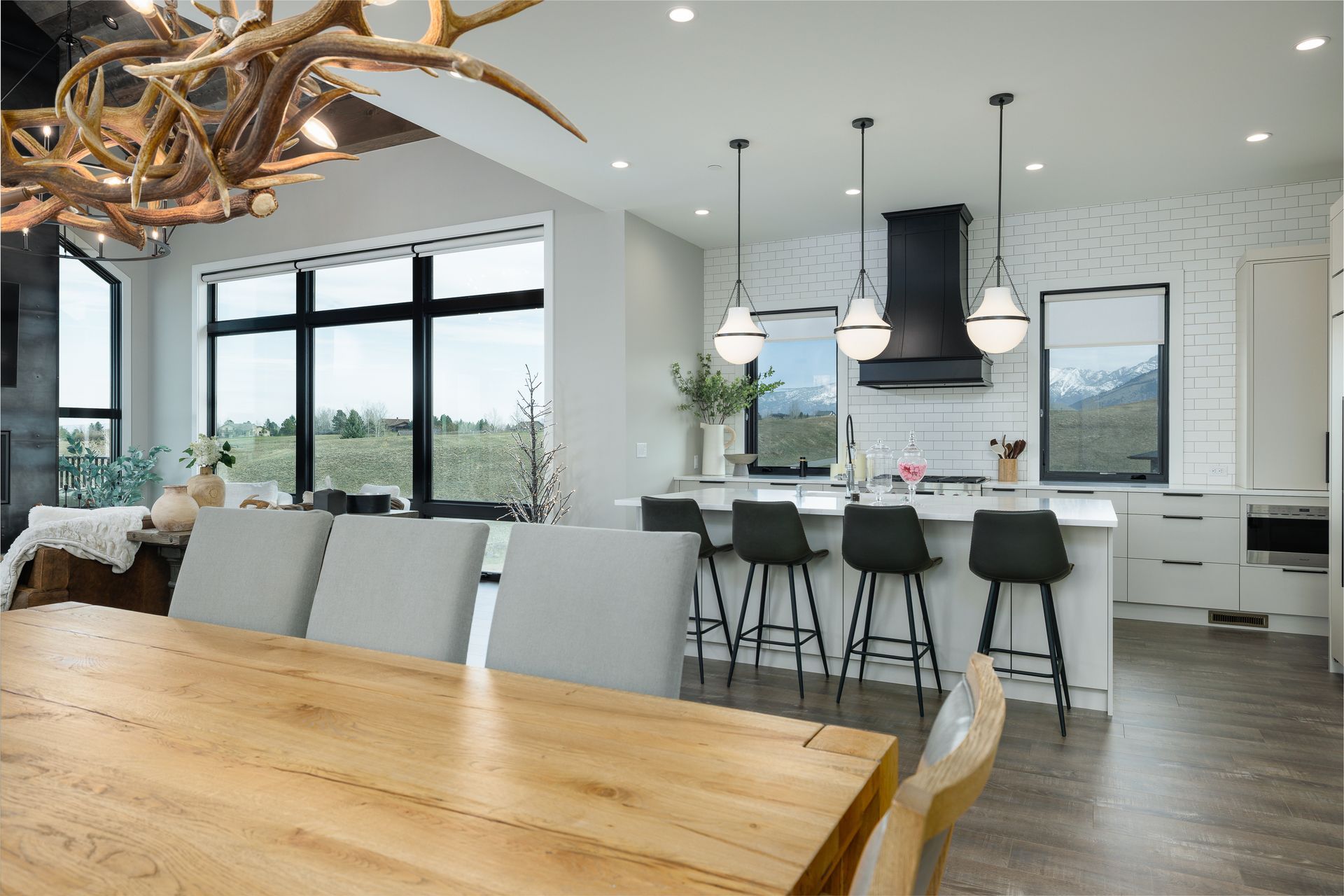 Modern open-plan kitchen and dining area with wood table, white cabinets, black stools, and large windows