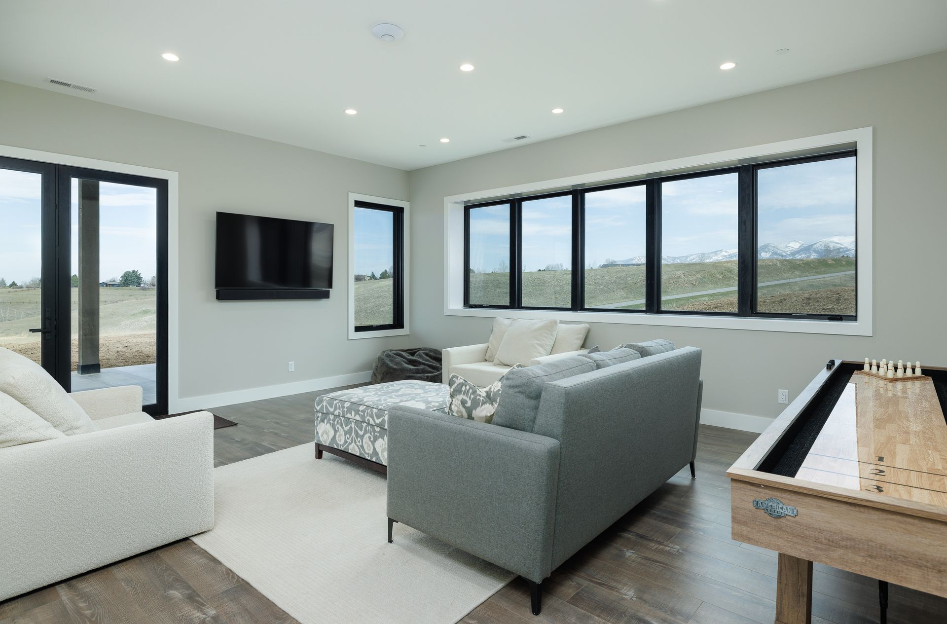 Modern living room with gray sofa, white chair, wall-mounted TV, and large windows overlooking a landscape