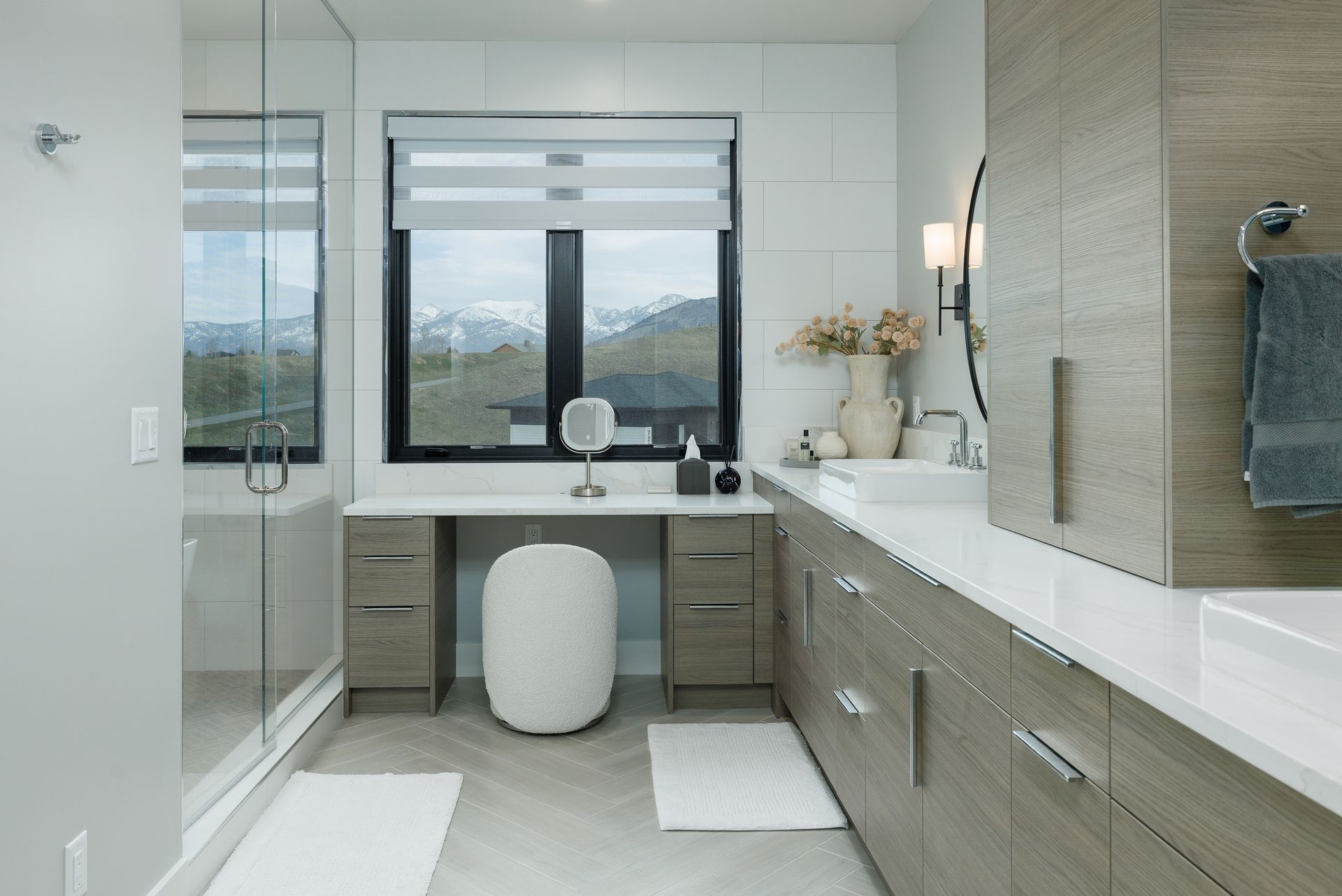 Modern bathroom vanity with large mirror, window, gray cabinetry, white counters, and a stool beneath the sink.