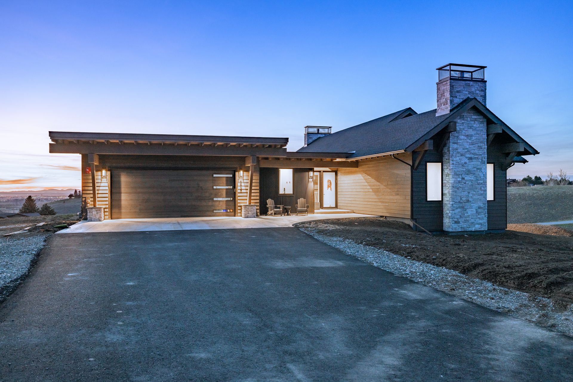 Modern house exterior at dusk with a stone chimney and attached garage on a paved driveway