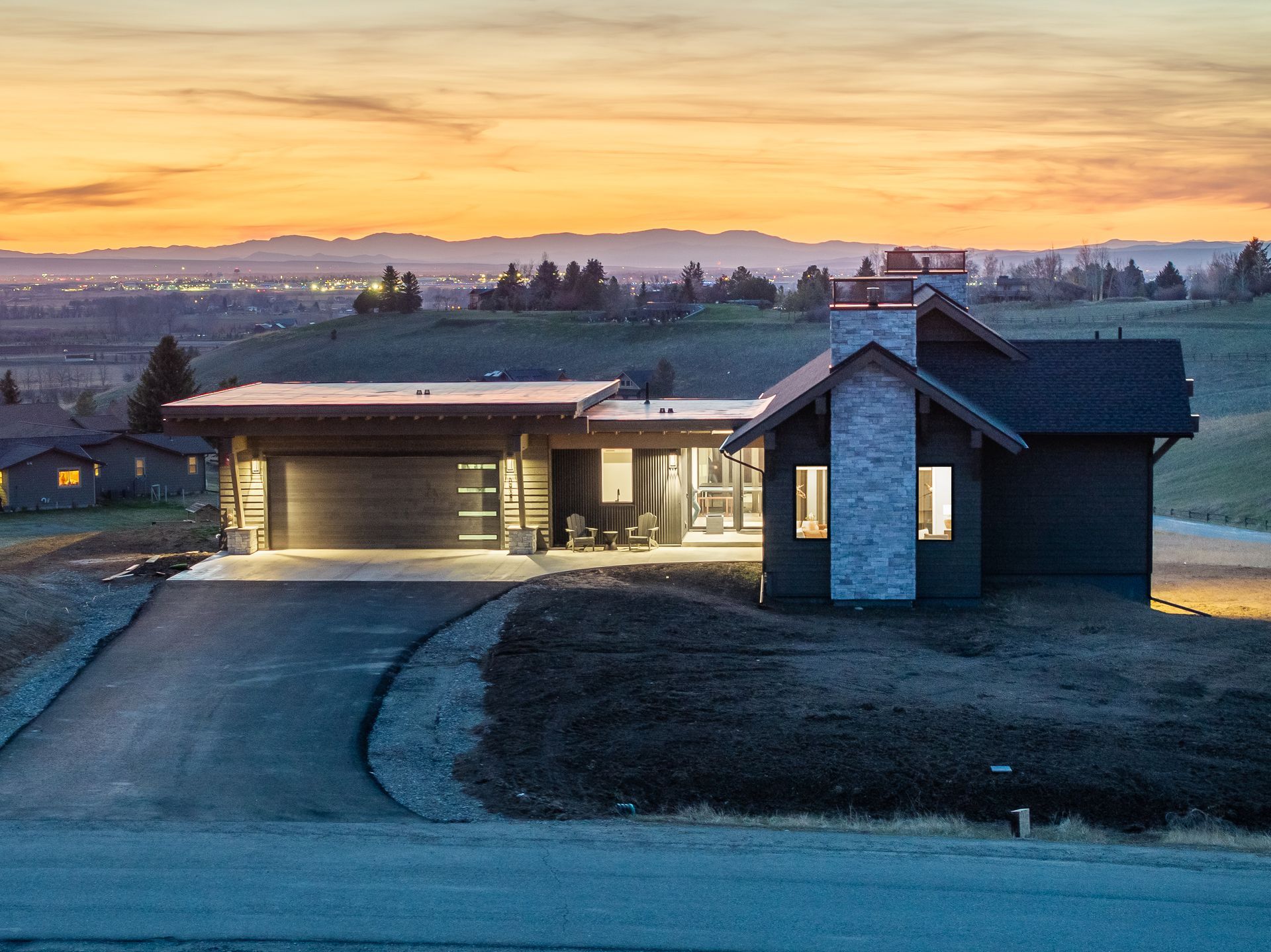 Modern house at dusk with warm lights, driveway, and rolling hills under a pink-orange sky