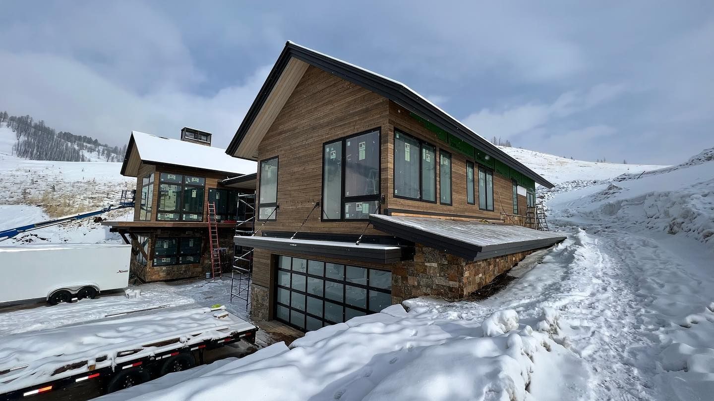 A modern wooden mountain home under construction, with large windows and a garage door, surrounded by deep snow.
