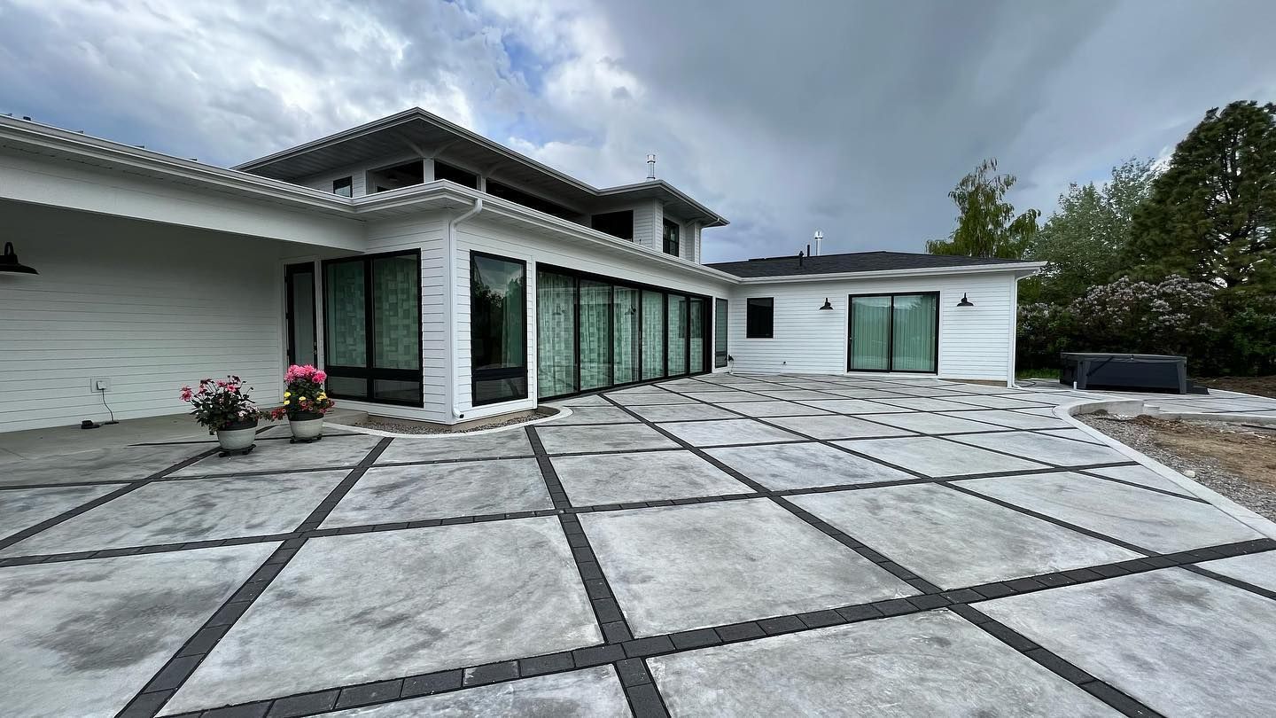 A white house with a large, modern patio featuring a diamond-patterned concrete floor under a cloudy sky.