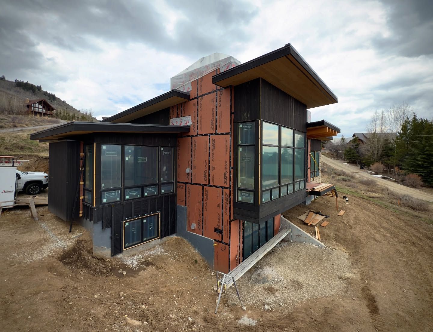 A modern house under construction with dark siding and large windows, framed by mountains and a dirt slope.