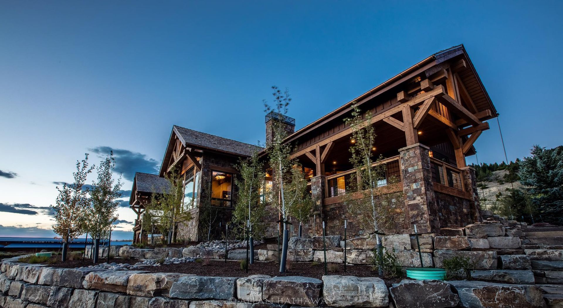 A rustic mountain home built with timber and stone, featuring a covered porch and stone landscaping at twilight.