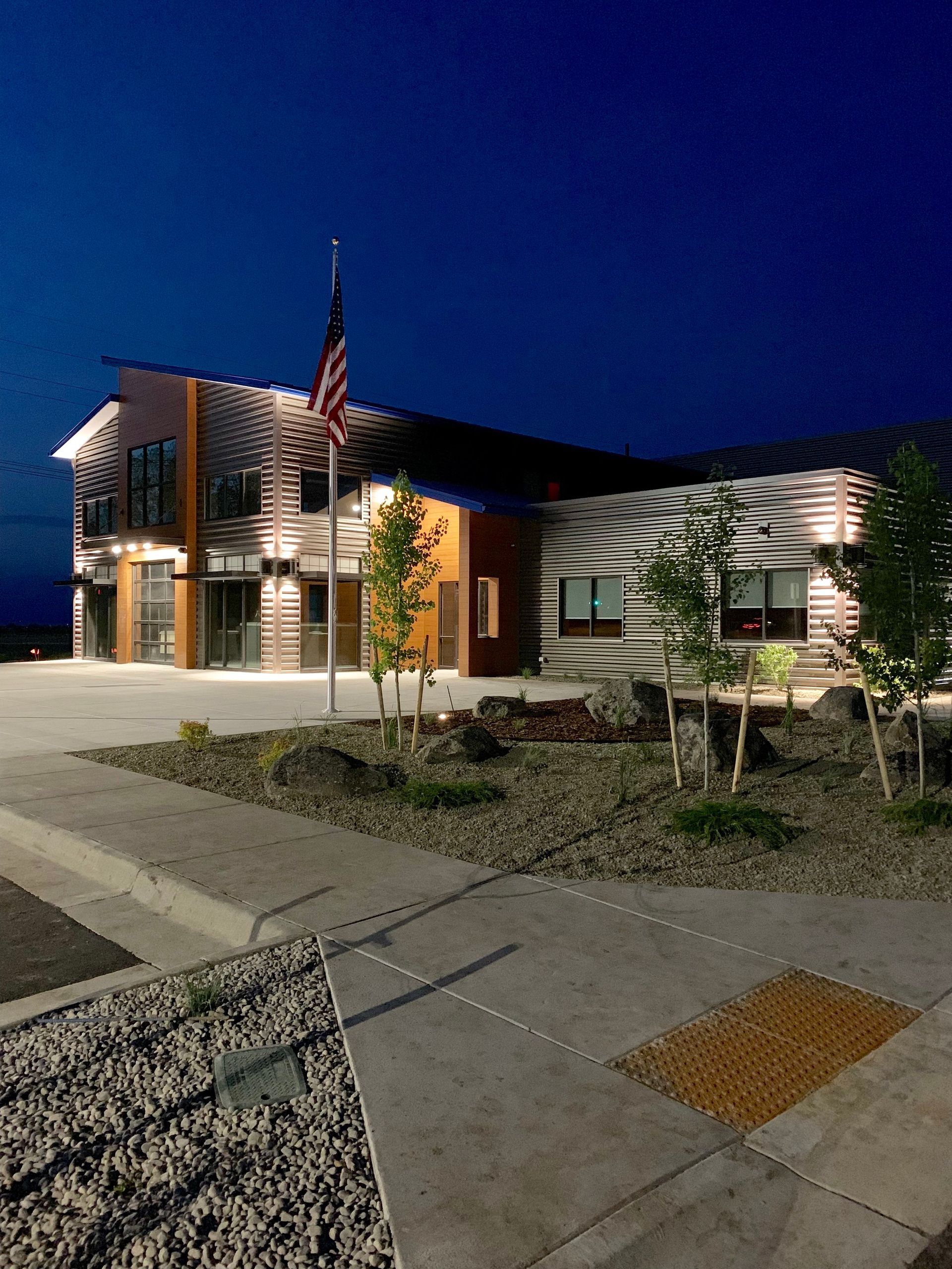 A modern building with metal siding and warm lighting at night, featuring a US flag and a landscaped front yard.
