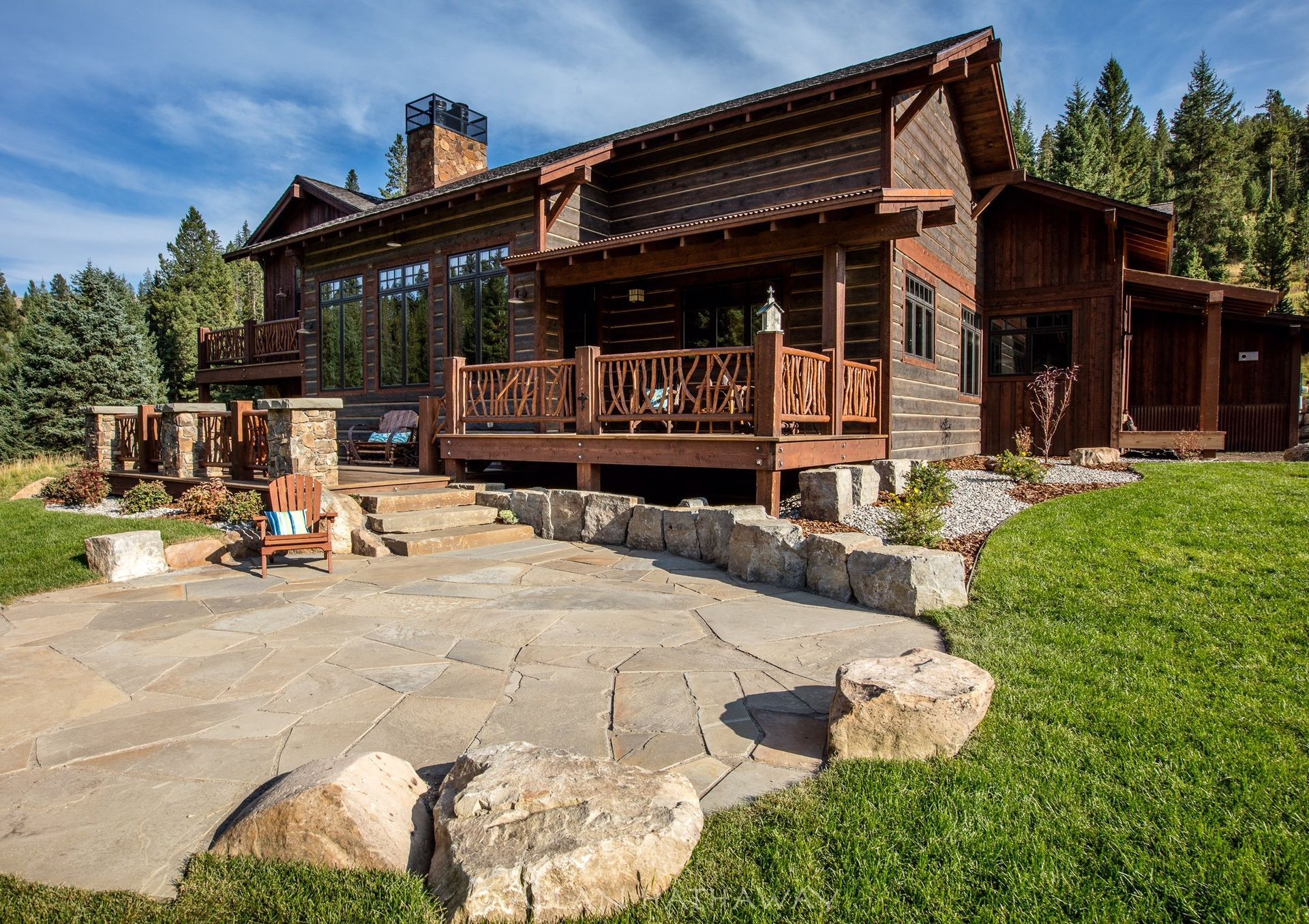 A rustic, multi-story log cabin with a stone patio, wooden deck, and chimney set against a forest background.