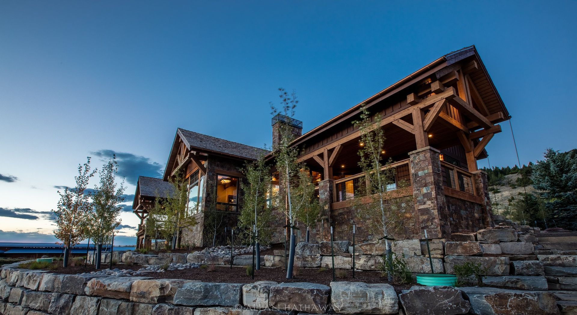 A rustic stone and timber lodge sits on a terraced hillside at twilight, illuminated by warm interior lights.