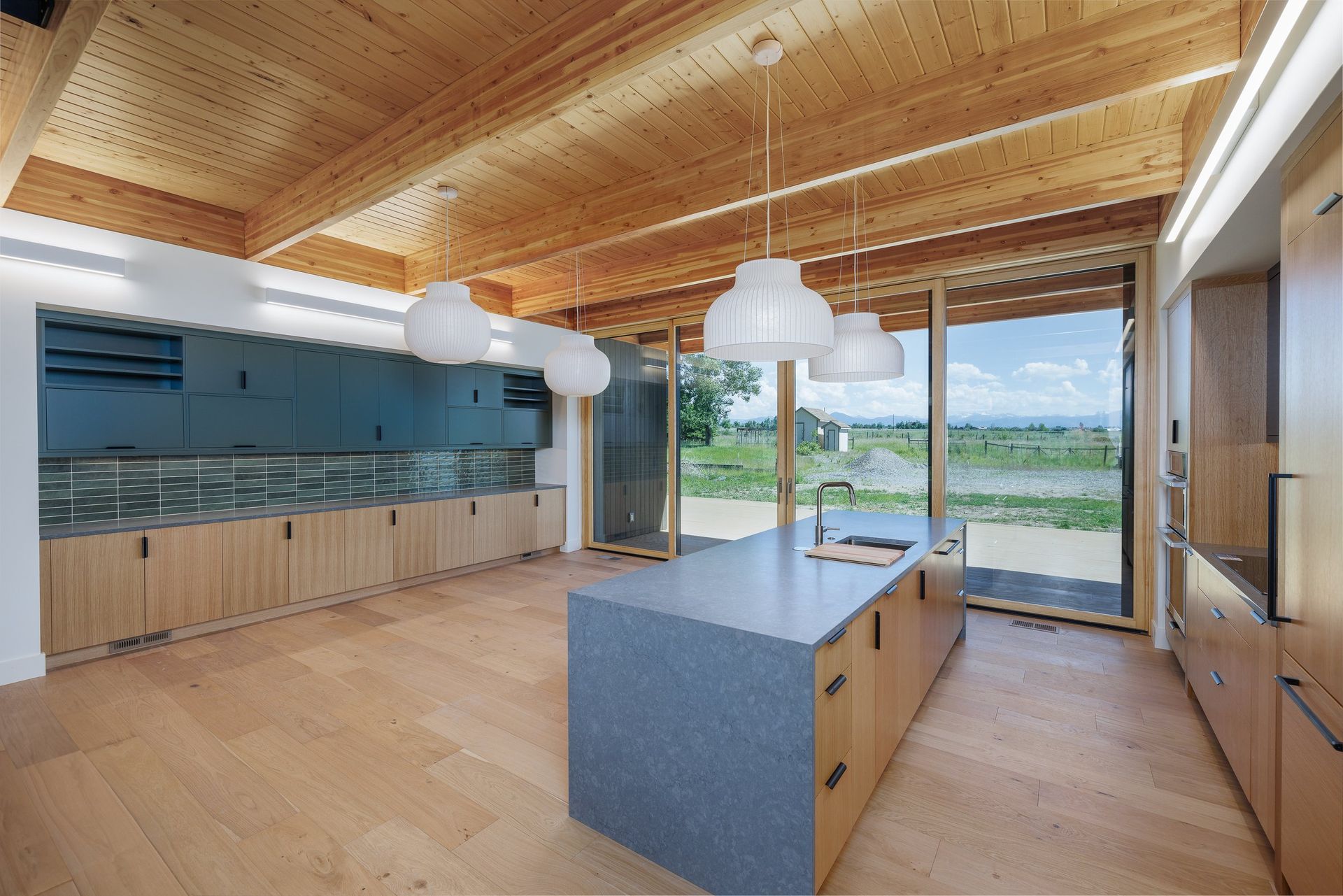 Modern kitchen with a gray island, wood cabinetry, pendant lights, and large glass doors looking out onto a field.