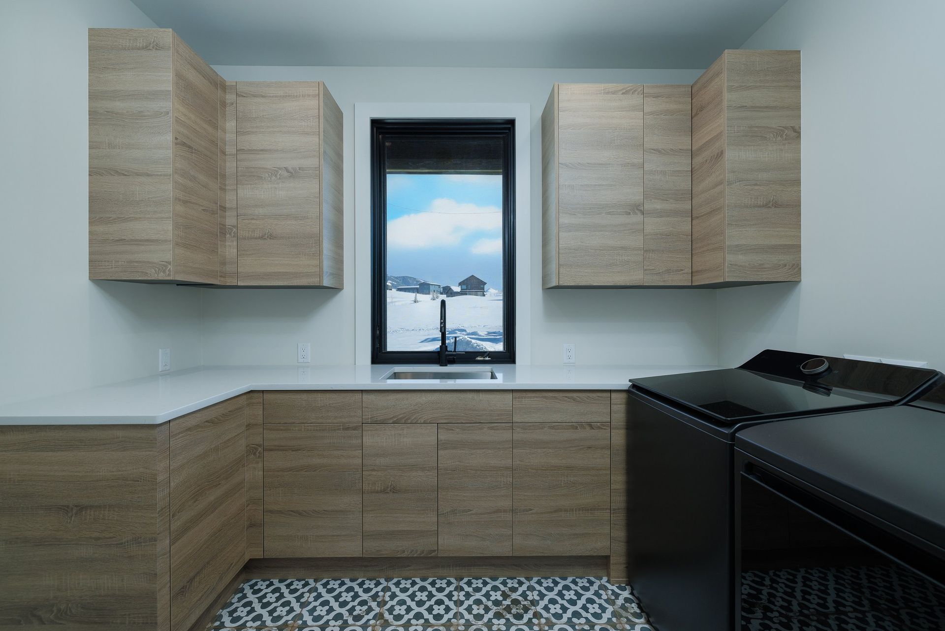 A laundry room with light wood cabinets, white countertops, a sink under a window, and black washer and dryer units.