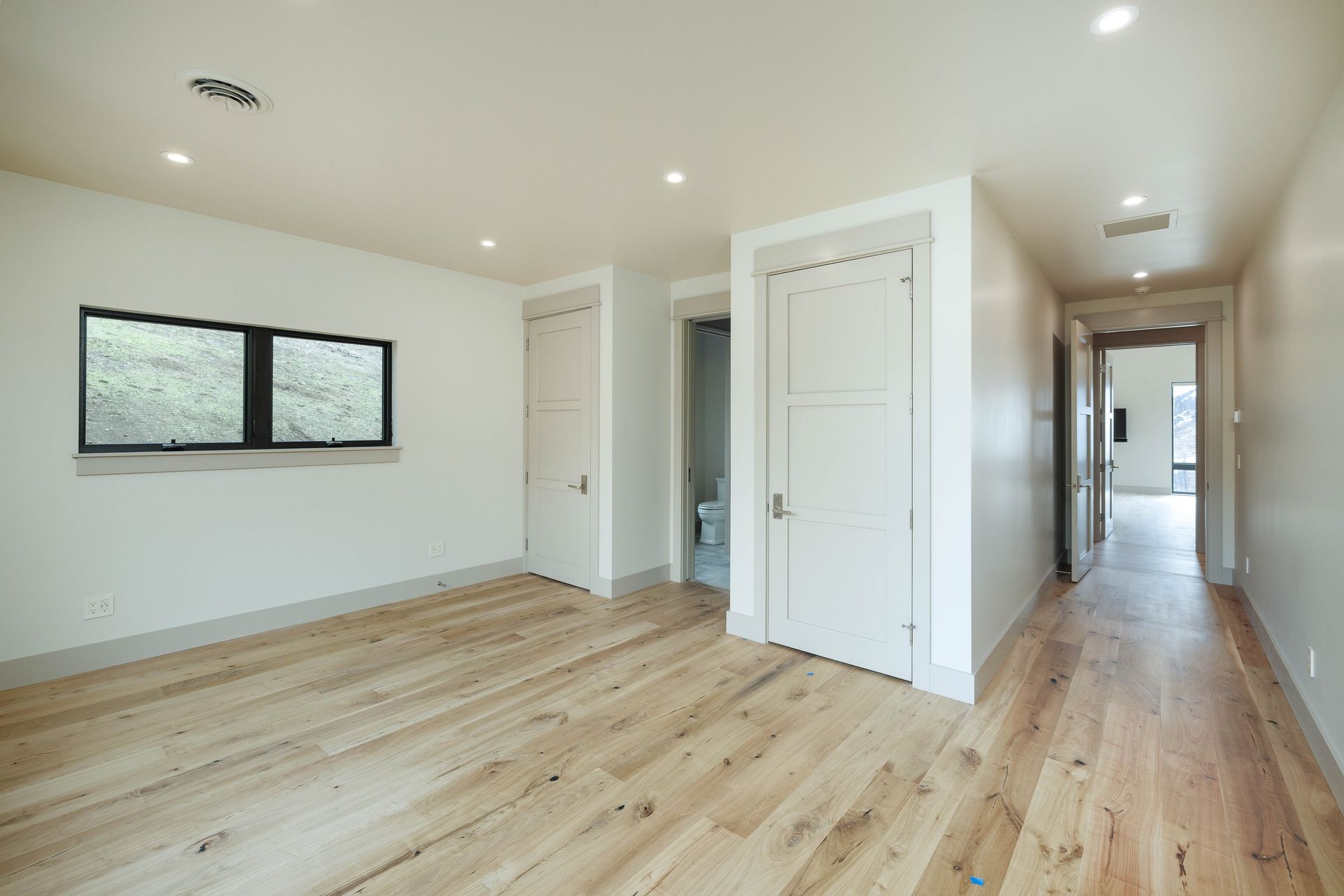 An empty room with light wood floors, white walls, a rectangular window, and two doorways leading to other spaces.