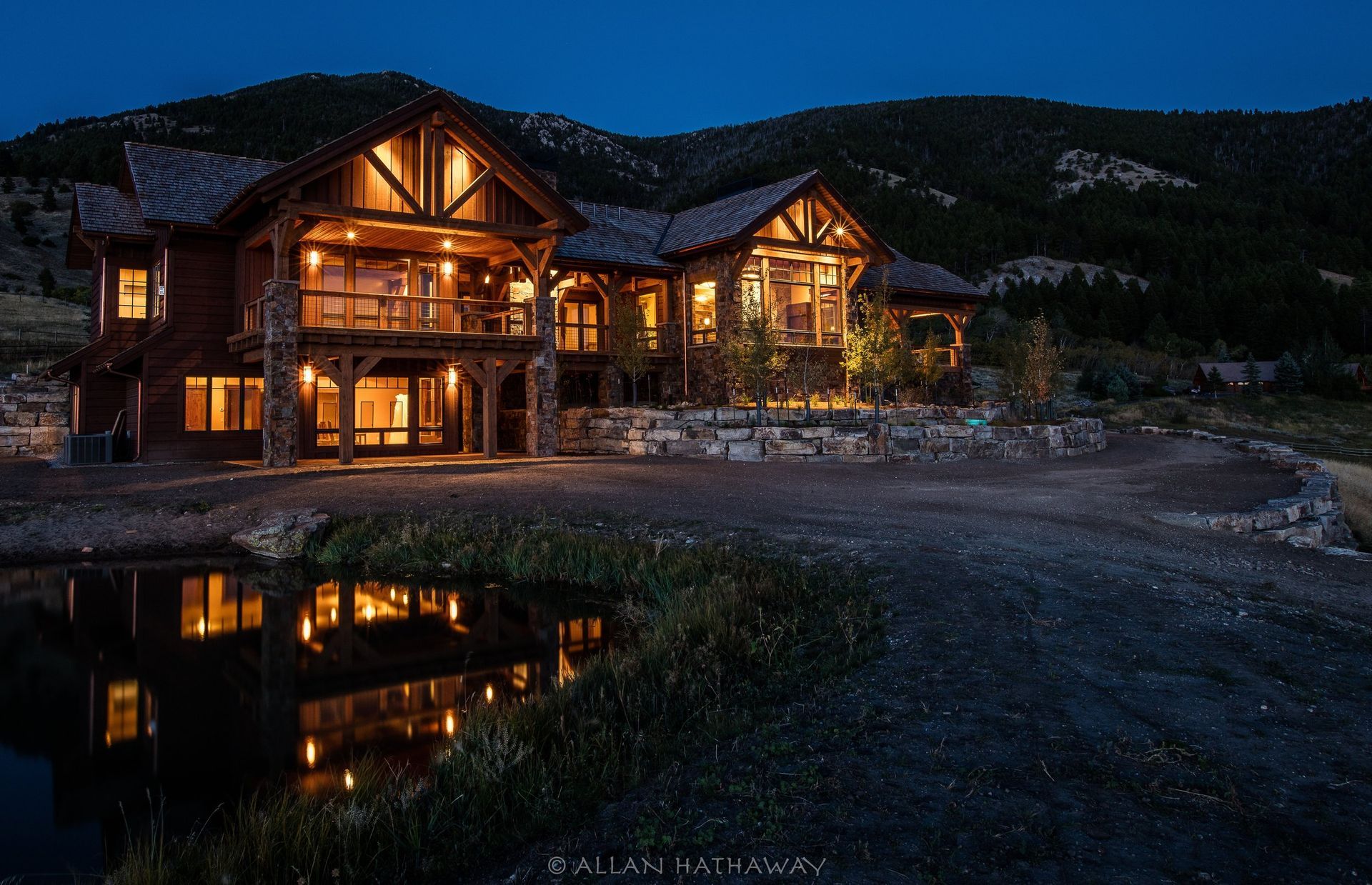 A multi-story timber-framed luxury mountain home glows warmly against a twilight sky, reflecting in a foreground pond.