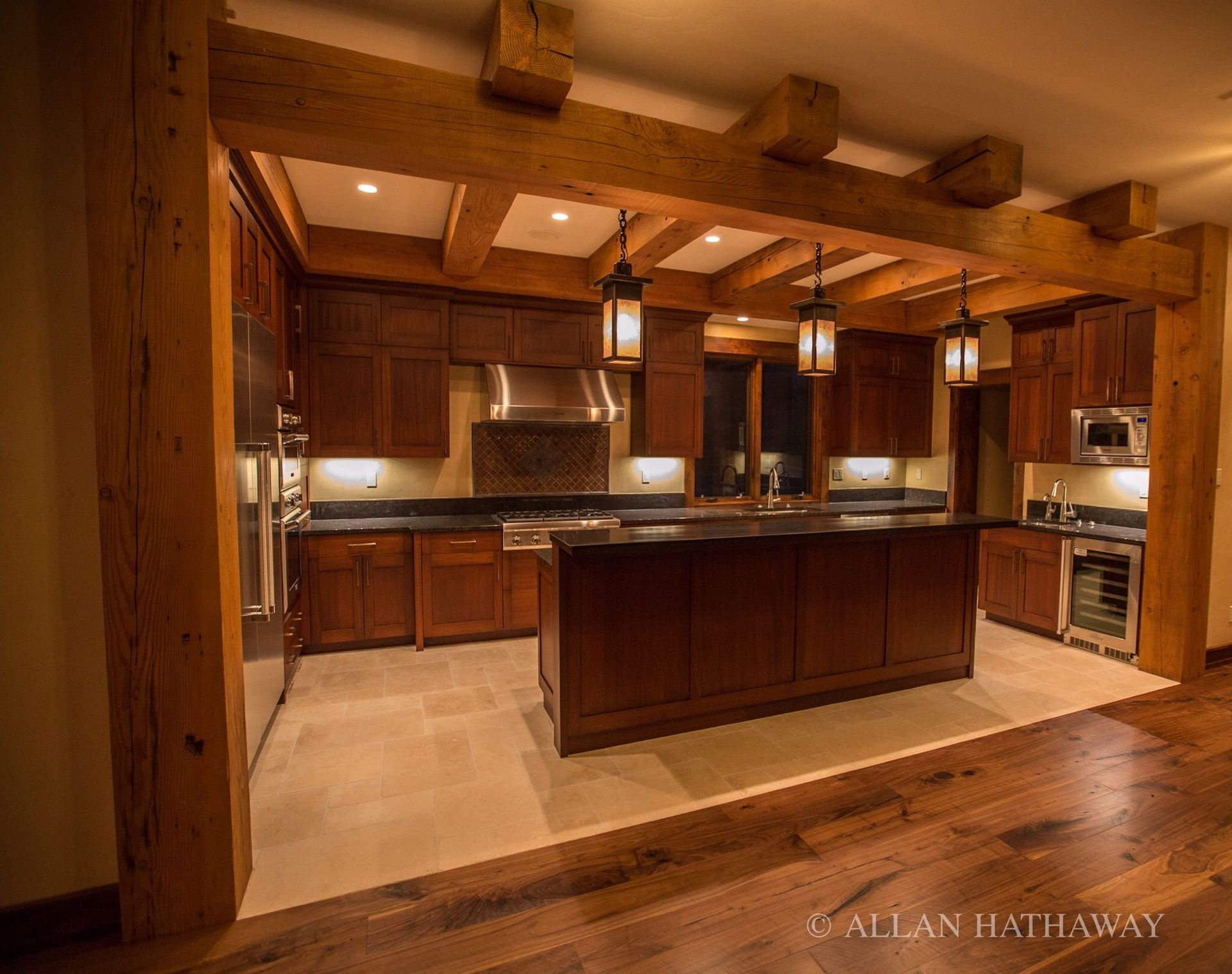 A modern kitchen featuring rich wood cabinetry, stone floors, dark countertops, and overhead exposed timber beams.