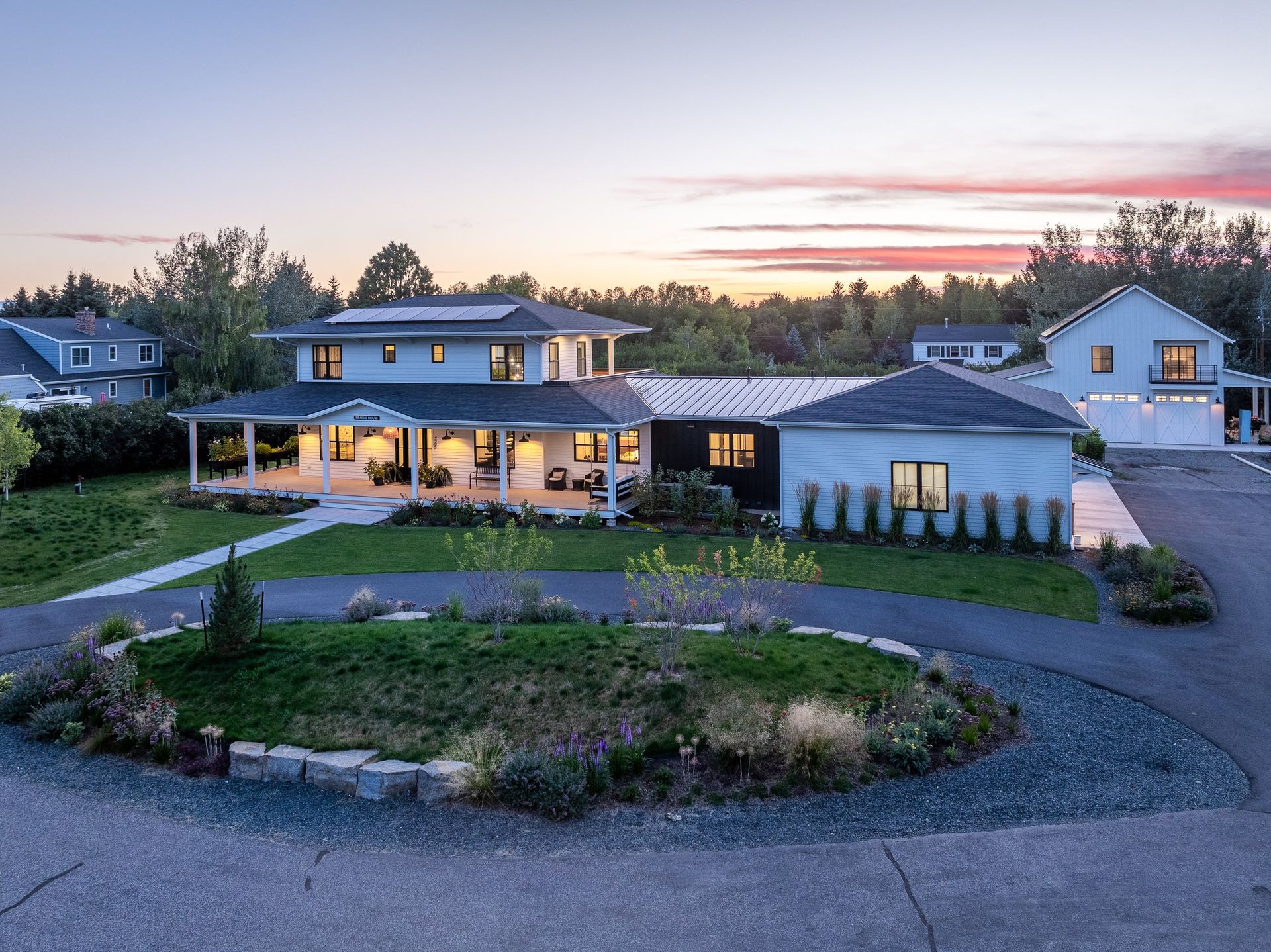 A two-story white house with a wrap-around porch and a circular gravel driveway at twilight.