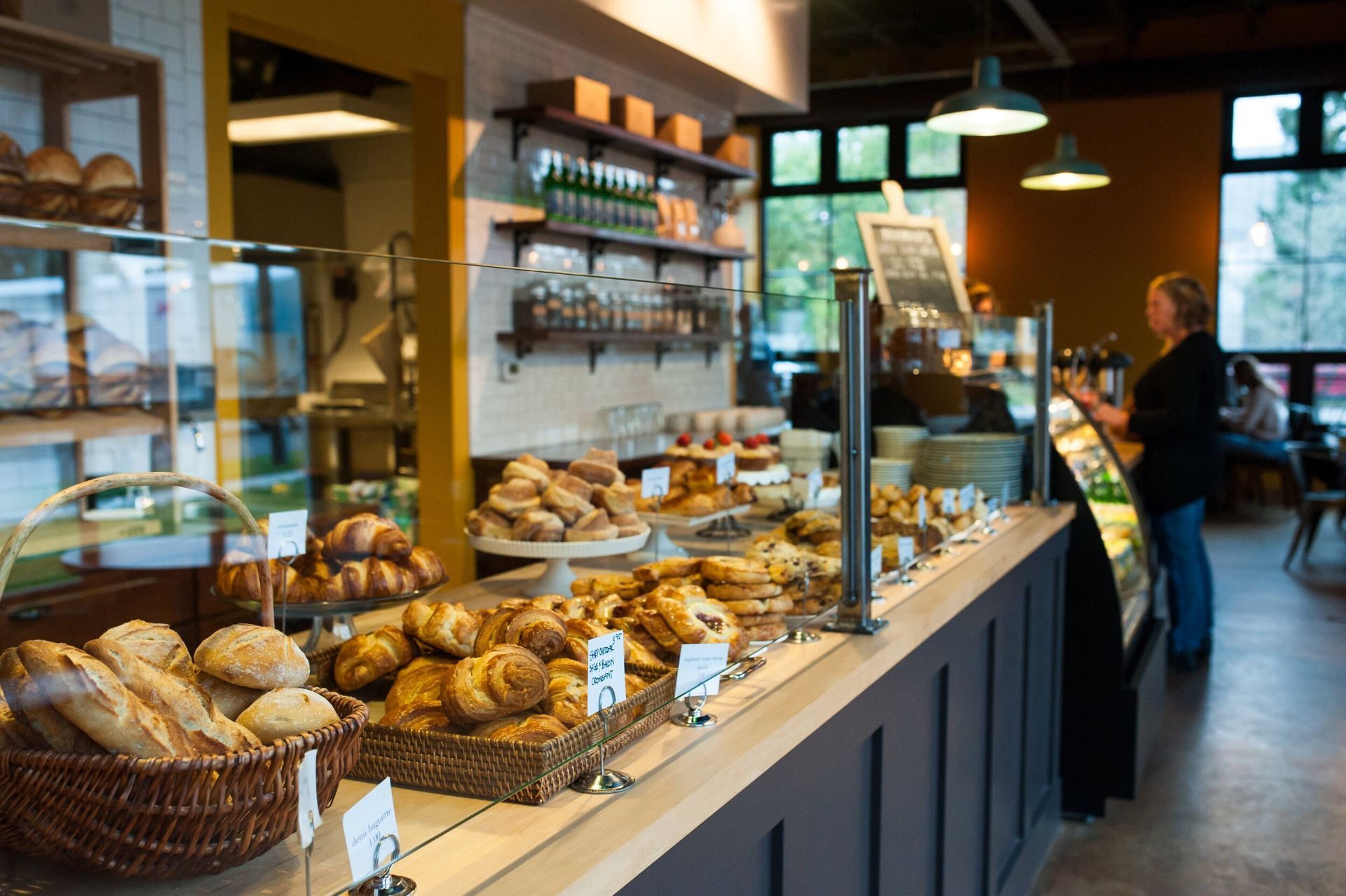 A bakery counter filled with various breads and pastries in baskets, with a staff member working in the background.