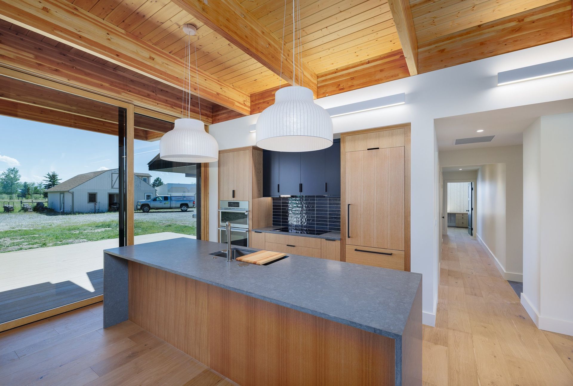 Modern kitchen with wood cabinetry, a large grey island, and pendant lights, opening to an outdoor view.
