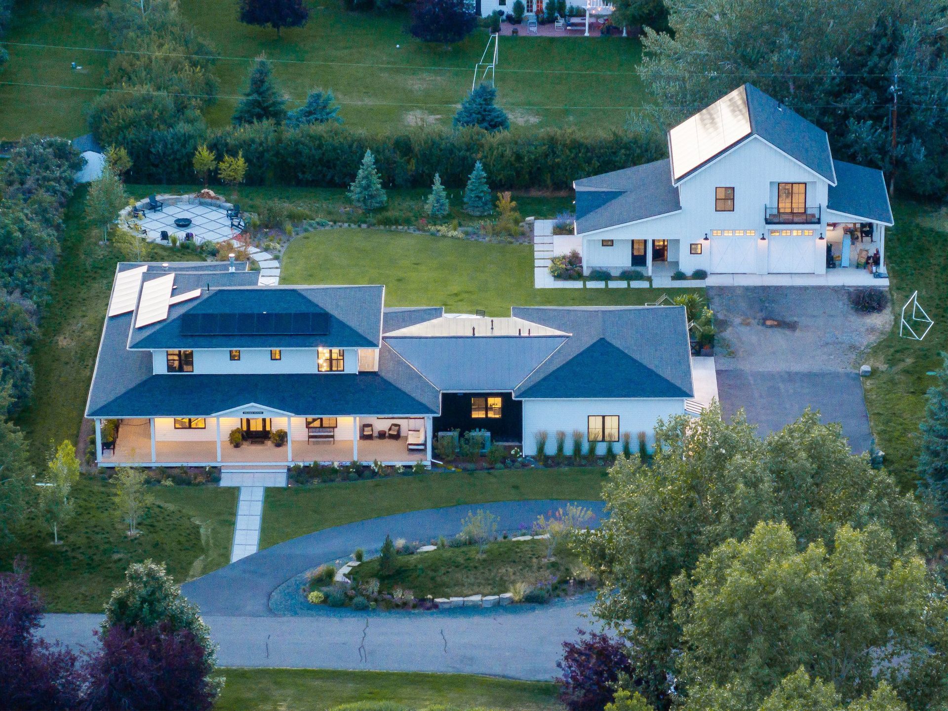 An aerial view of a white, multi-building residential property with dark roofs, green lawns, and trees at dusk.