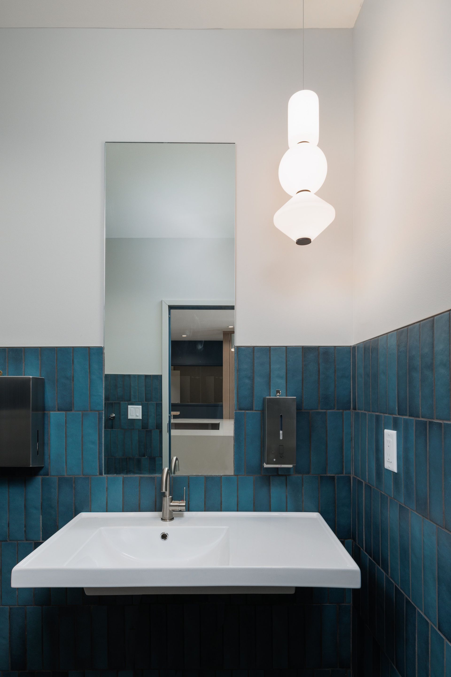 A white wall-mounted sink set against dark blue tiled walls, featuring a rectangular mirror and a modern pendant light.