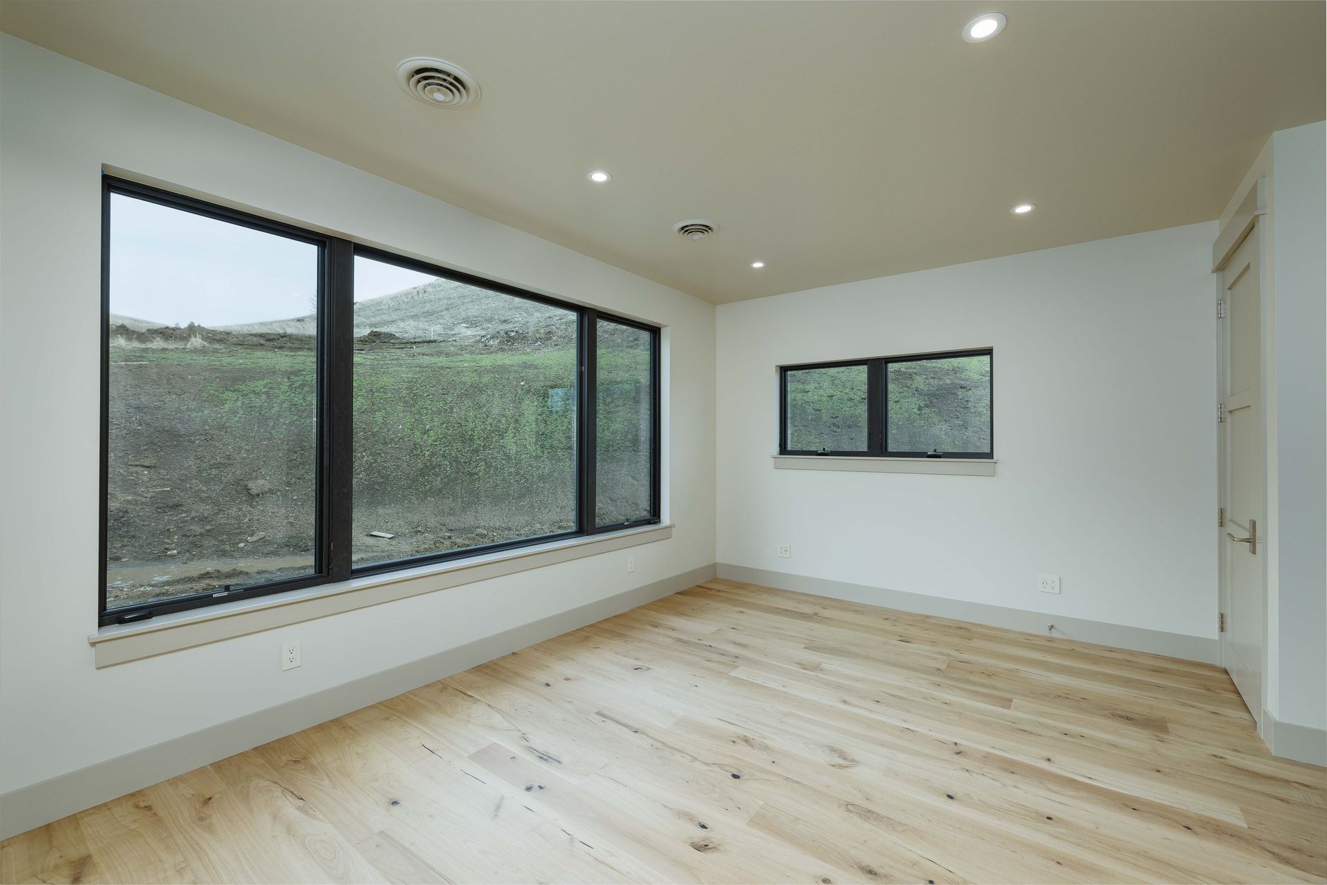 A brightly lit, empty room featuring light wood flooring, white walls, and a large window looking out onto a grassy hill.