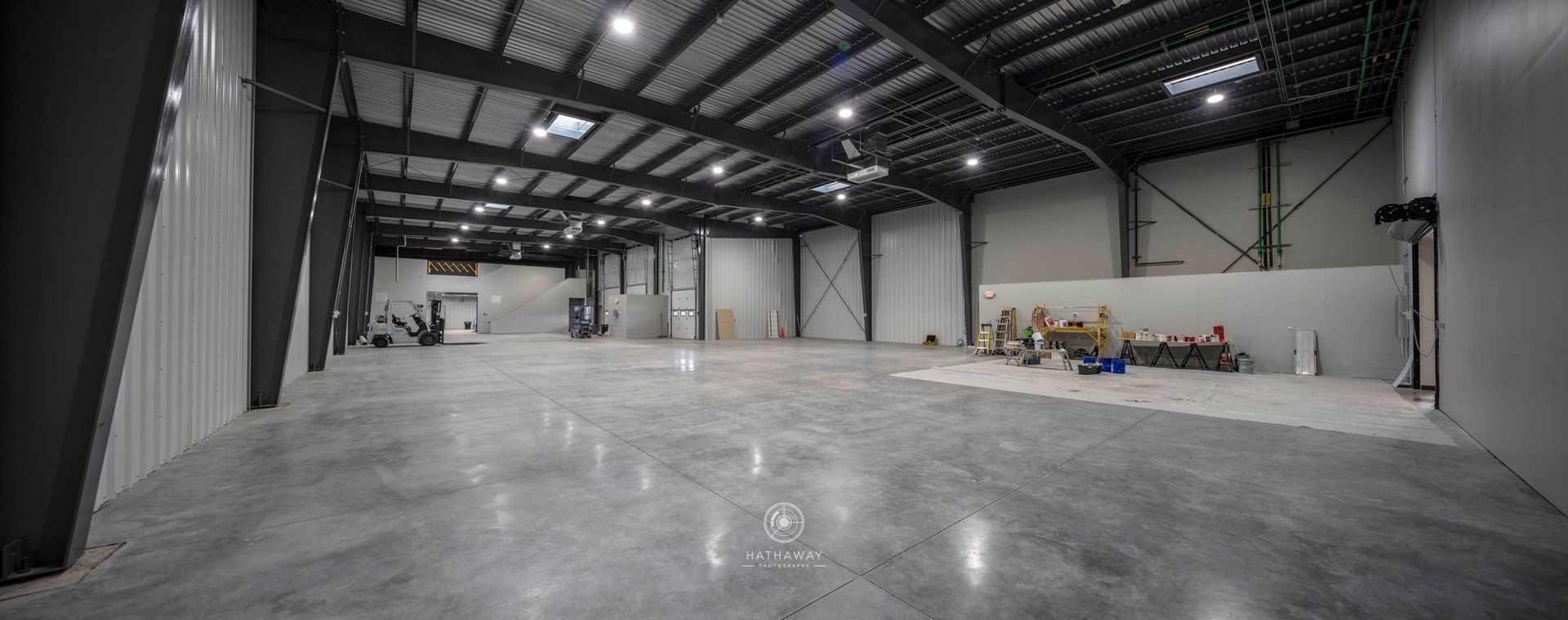 A wide, empty industrial warehouse with gray concrete floors, white walls, and a high, dark steel-beamed ceiling.