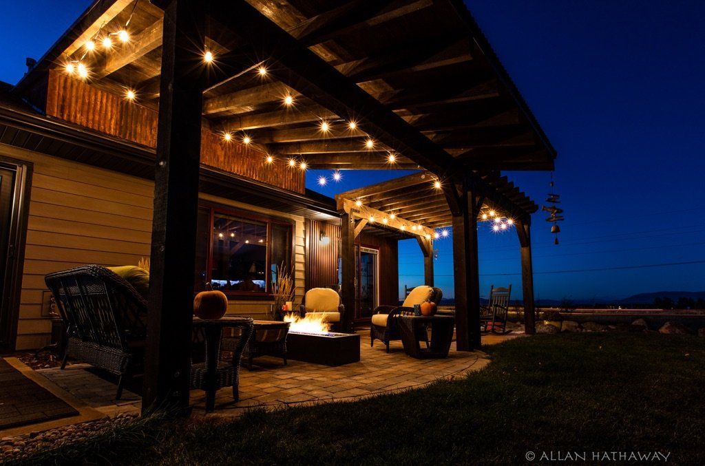 A cozy patio at dusk, illuminated by string lights with comfortable outdoor seating and a fire pit under a wood pergola.