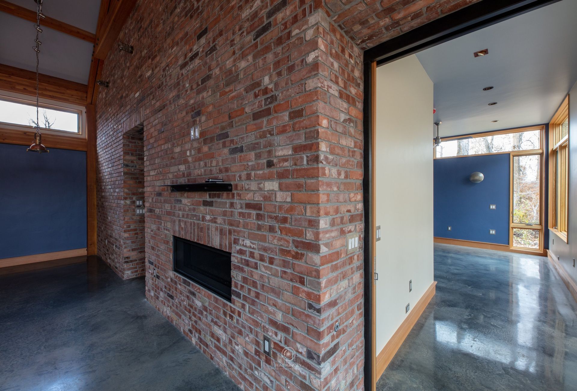 A central brick fireplace wall separates two rooms with blue accent walls, concrete floors, and large windows.