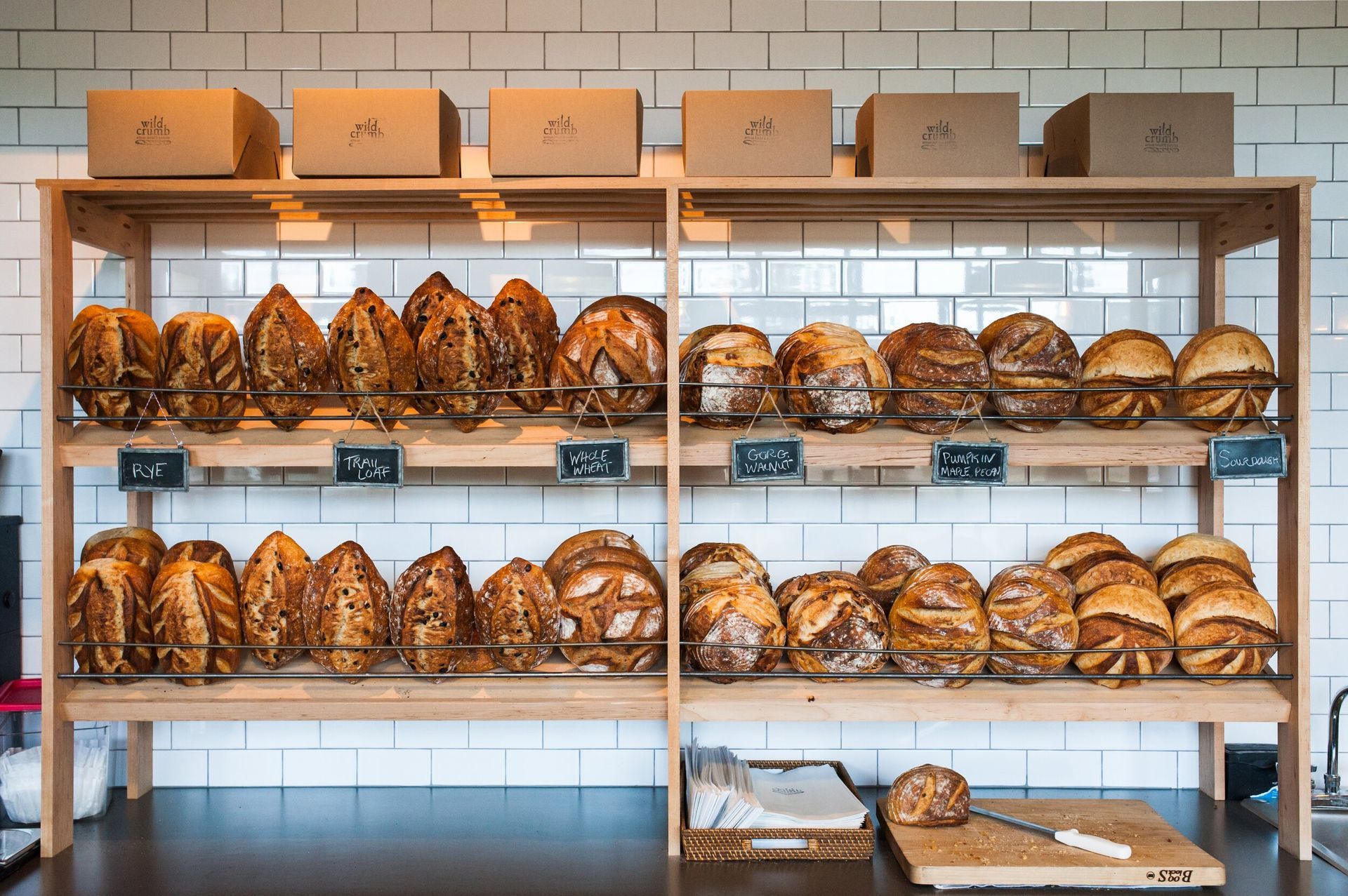 A wooden shelving unit filled with various crusty artisan loaves of bread in front of a white tiled wall.