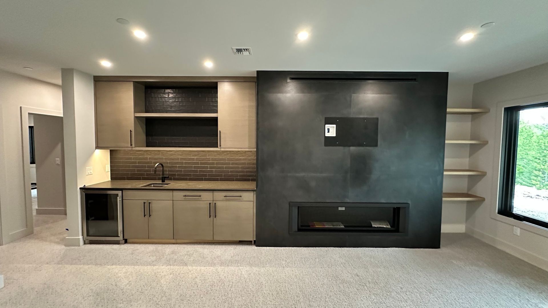 A wet bar with tan cabinets and a dark tile wall featuring an integrated TV and linear fireplace next to open shelving.