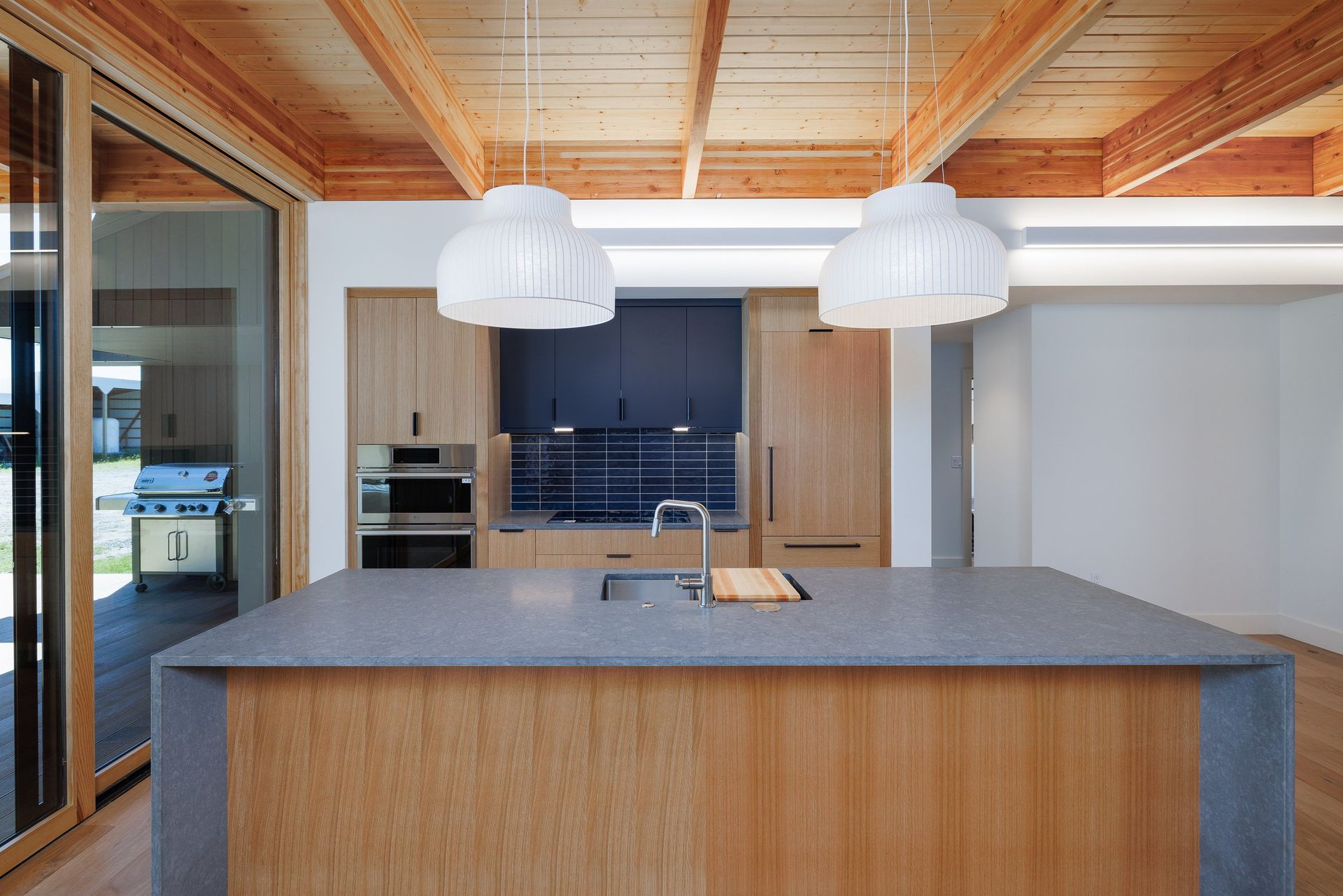 A modern kitchen with a wooden ceiling, light wood cabinets, dark blue backsplash, and a central island with a grey top.