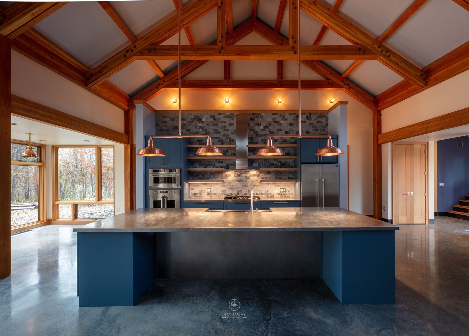 Modern kitchen with a large blue island, metal countertops, and copper pendant lights under an exposed wood-truss ceiling.