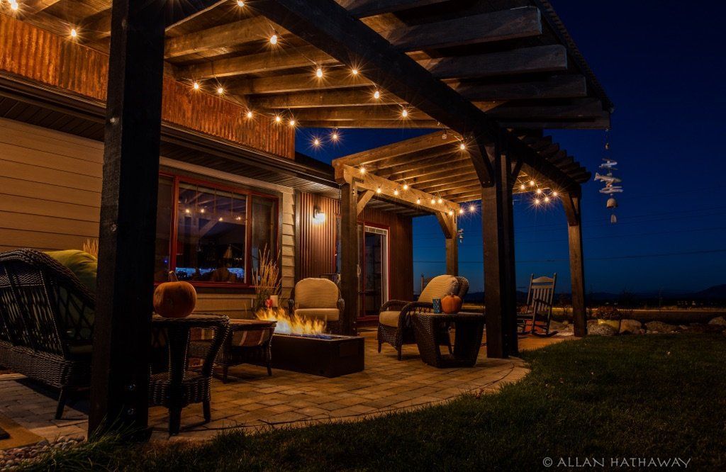 A glowing patio fire pit and seating area under a wooden pergola with string lights against a deep blue twilight sky.