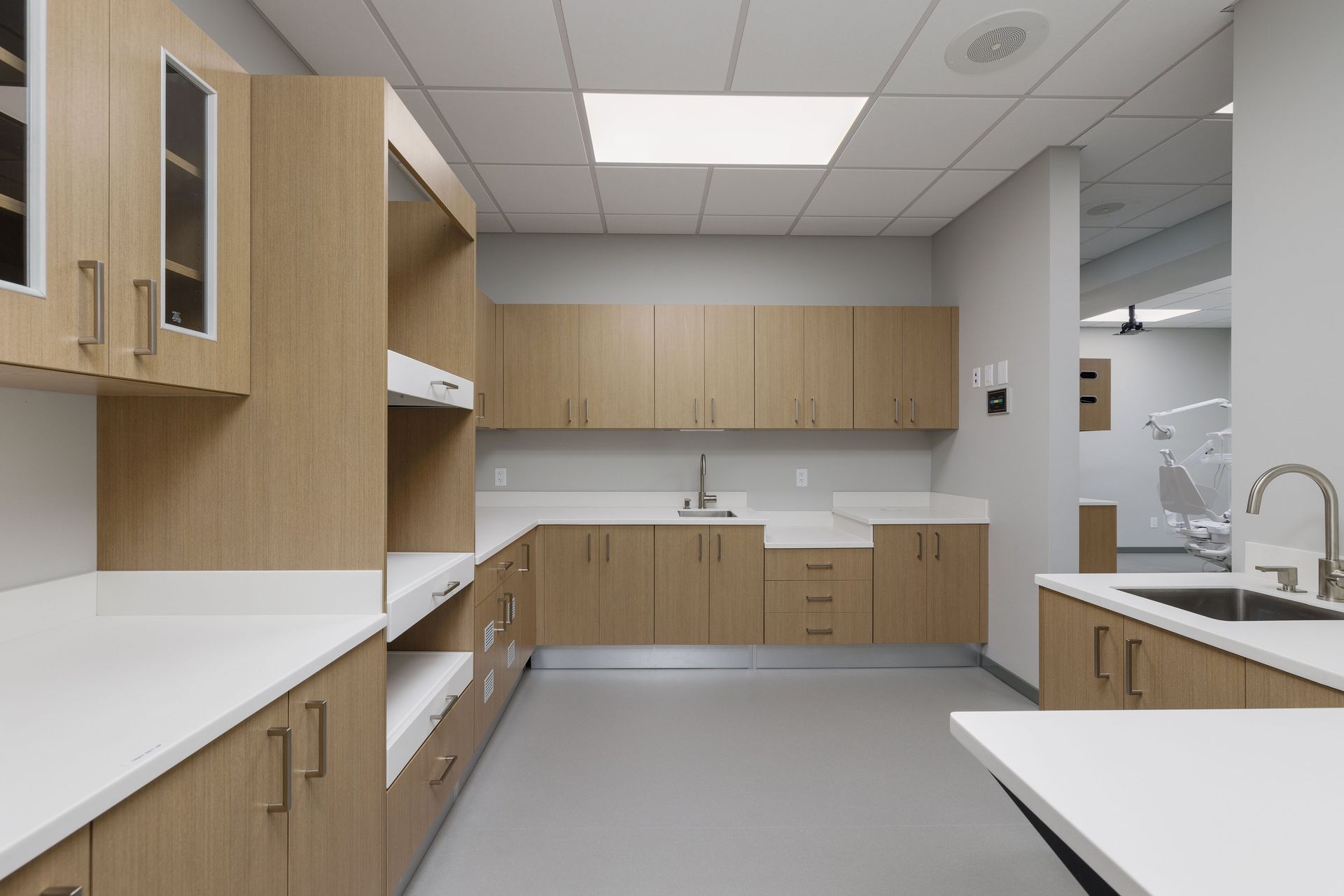 A modern dental sterilization room featuring light wood cabinets, white countertops, a sink, and a view into a clinic.