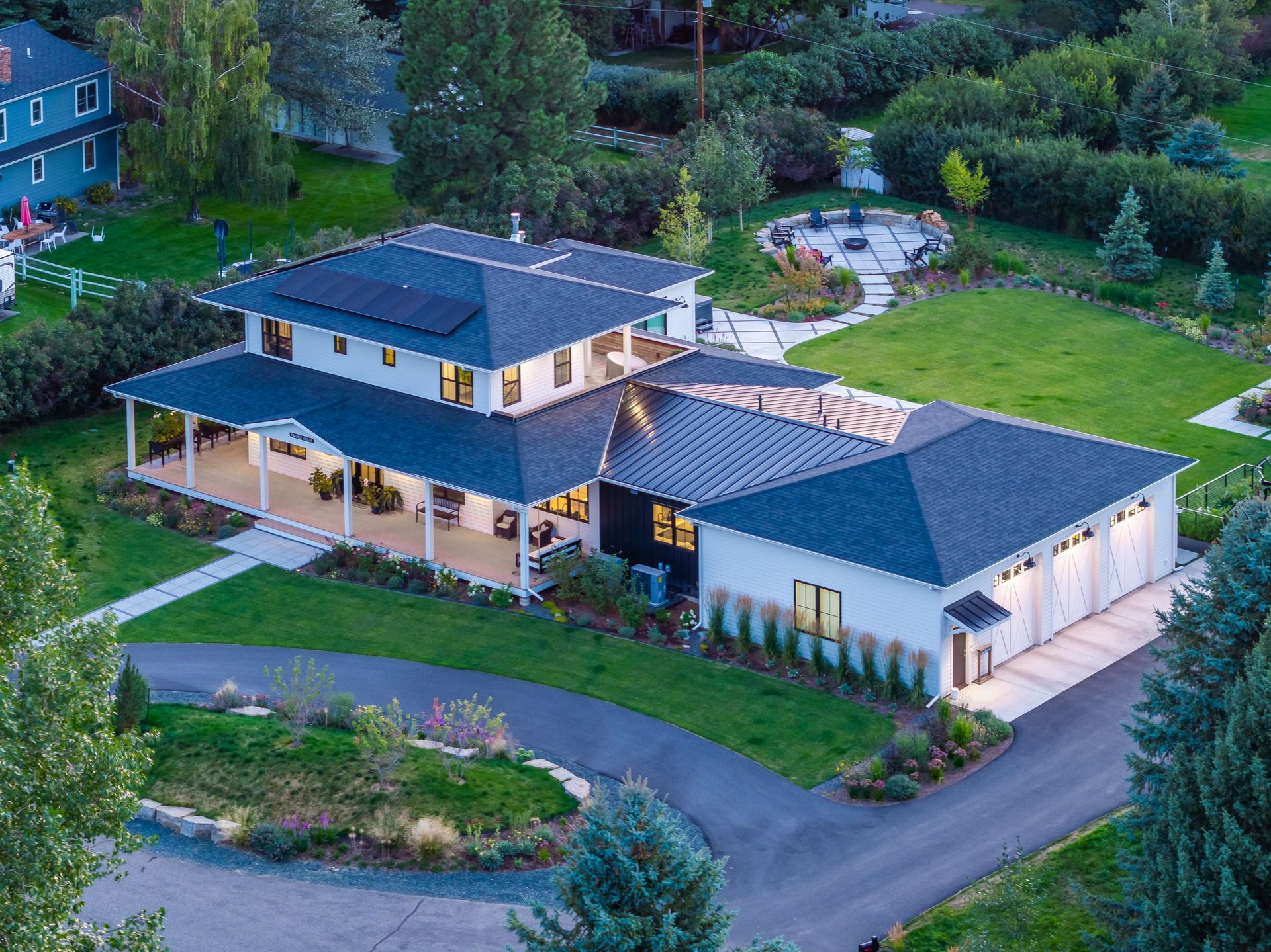 Aerial view of a white, two-story modern farmhouse with a dark shingle roof, a wrap-around porch, and a three-car garage.