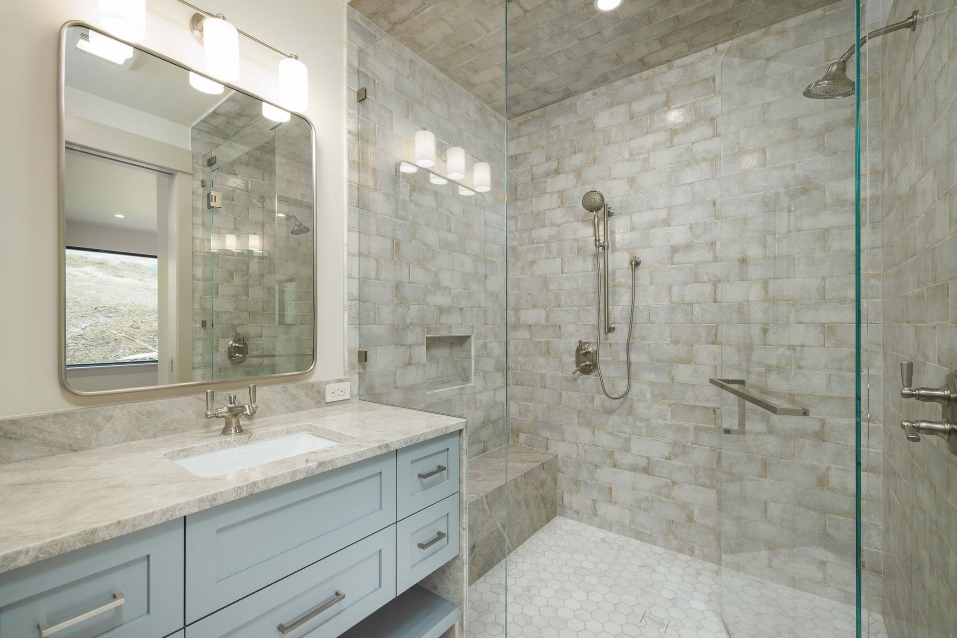 A modern bathroom with a light blue vanity, marble countertop, mirror, and a glass-enclosed shower with stone tile walls.
