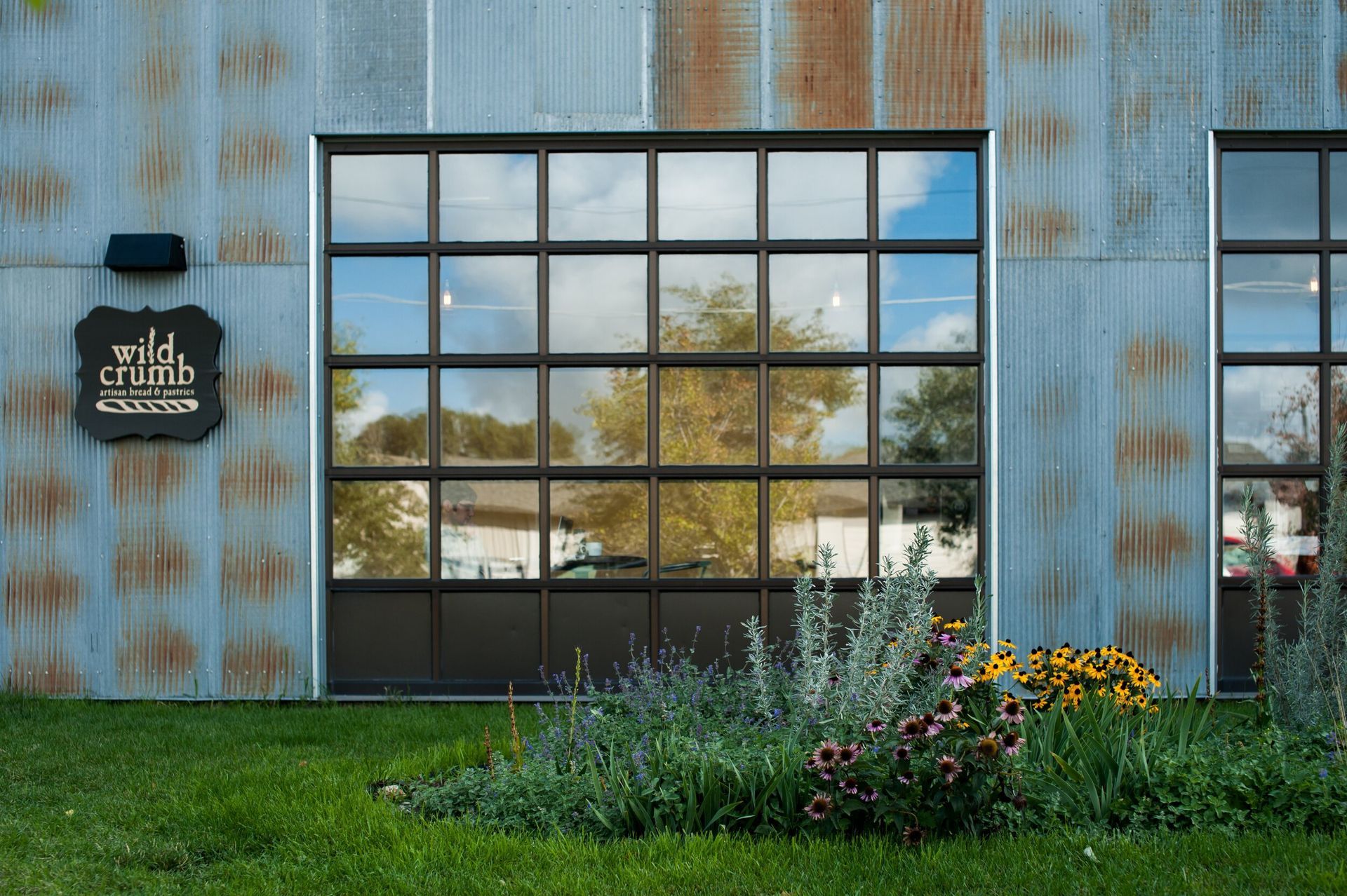 The Wild Crumb bakery storefront features a weathered metal exterior, large grid-pane windows, and a patch of garden.